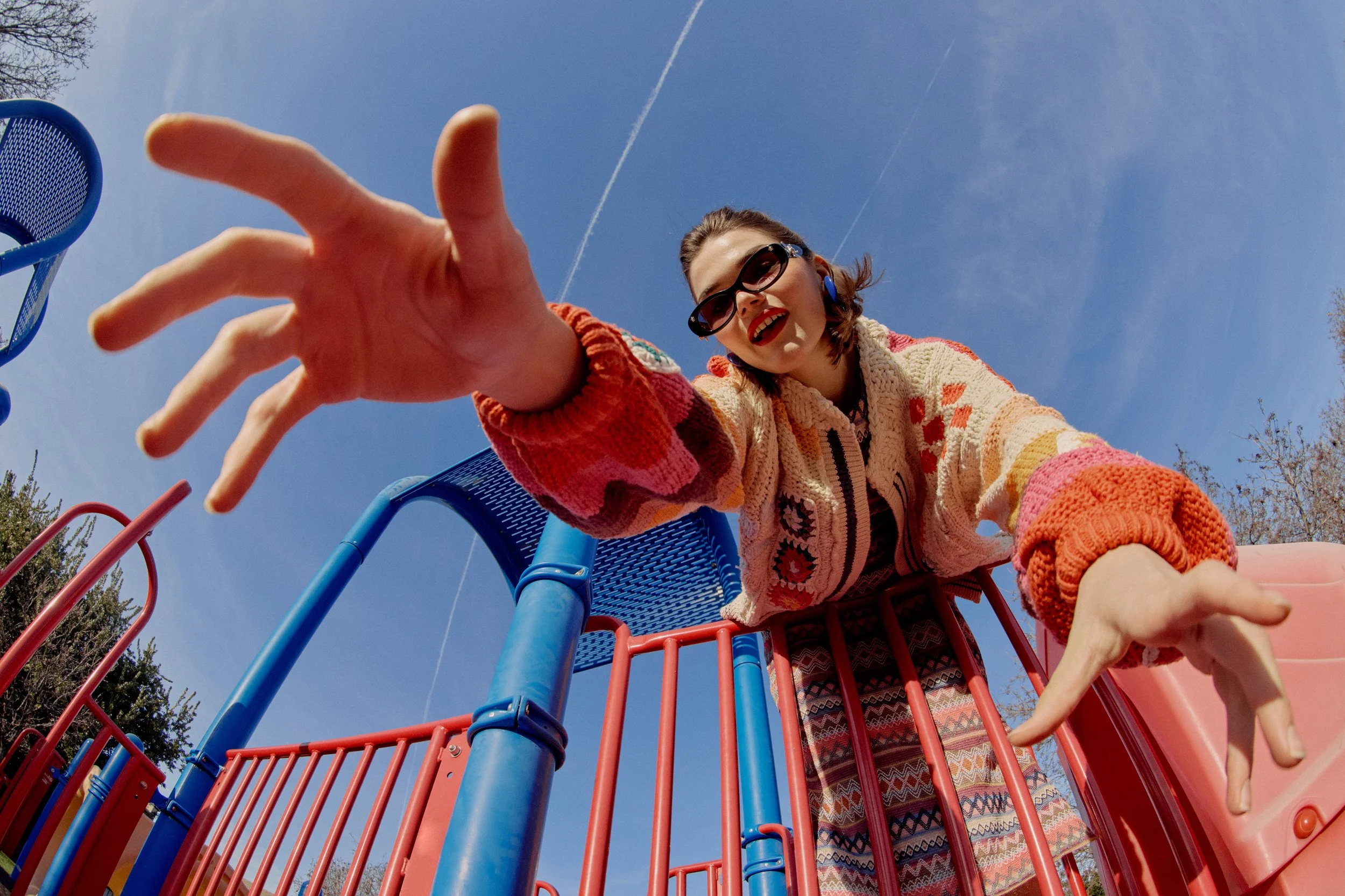 A woman with dark hair, wearing sunglasses, a colorful knit sweater, and patterned pants, is smiling and reaching toward the camera while standing on a blue and red playground slide outdoors on a sunny day.