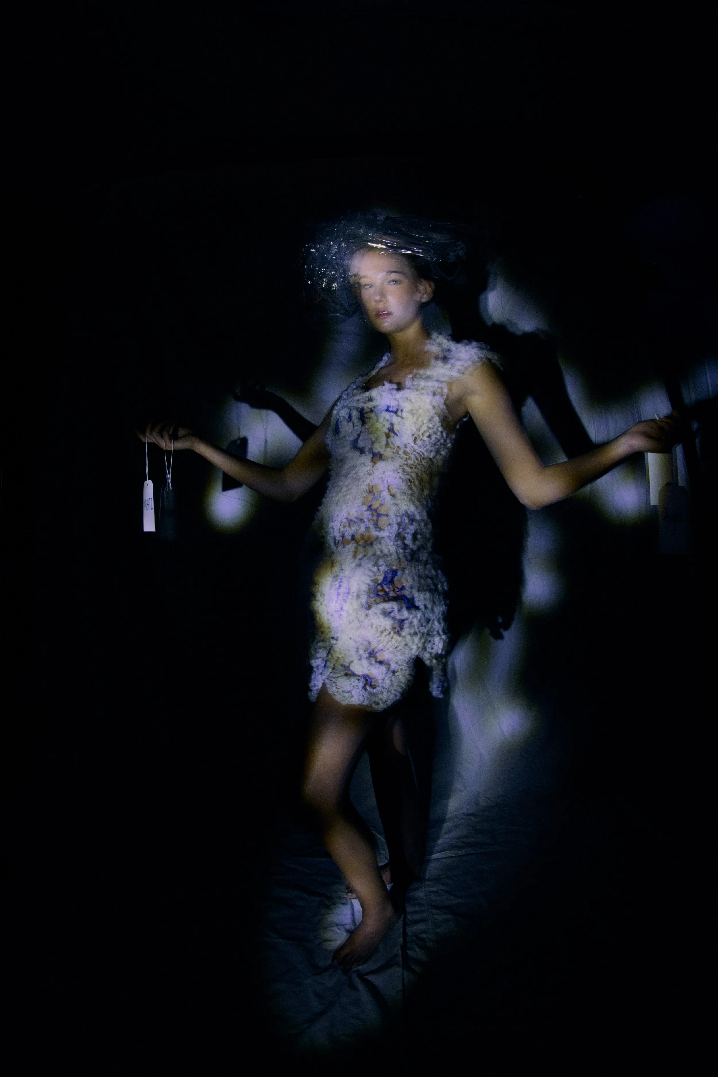A woman lying in a dark room with a reflective surface on her head, wearing a lacy dress, with other people partially visible around her in a dimly lit setting.