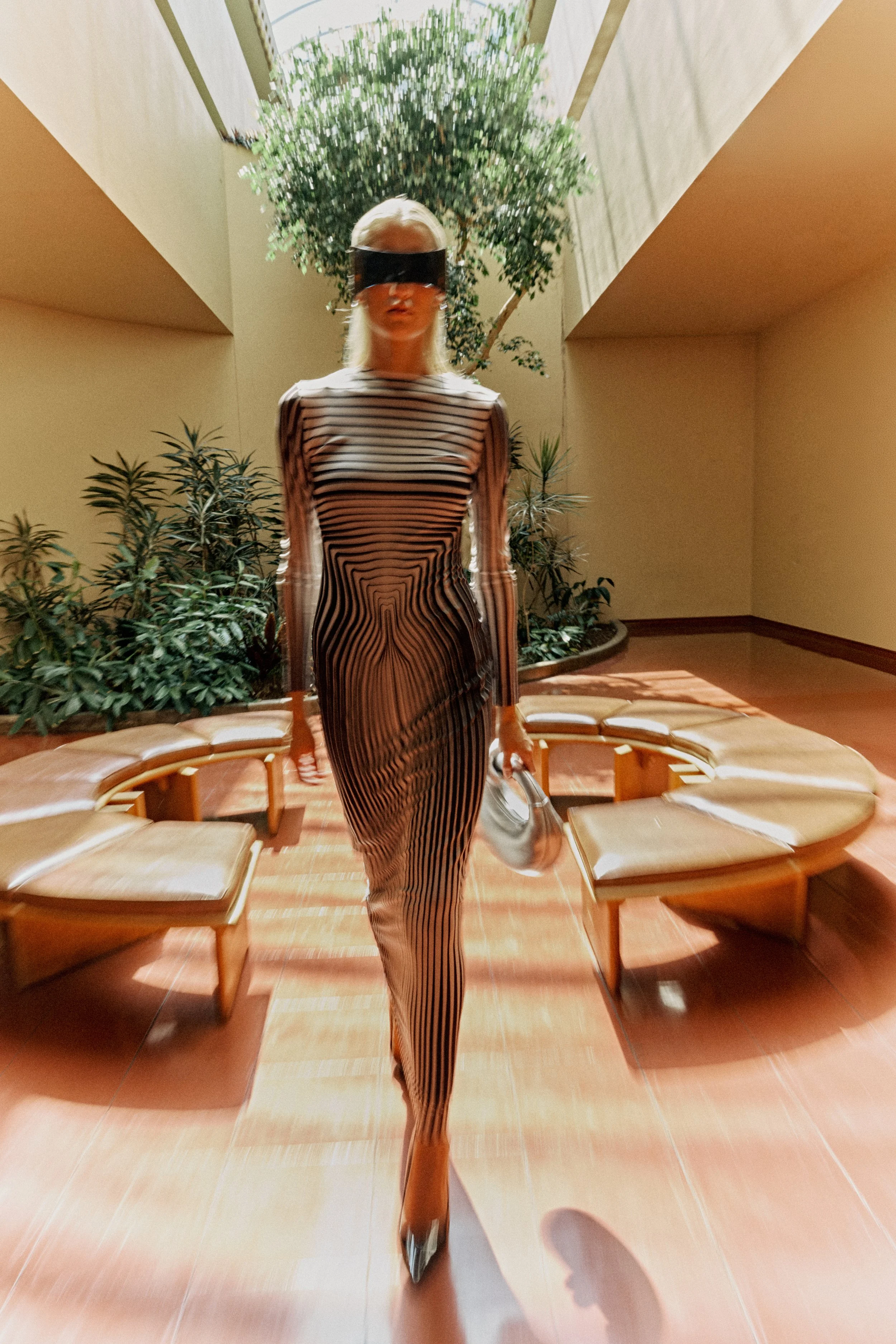 A woman wearing a striped dress and high heels walking in a room with benches and green plants, with a large plant and natural light overhead.