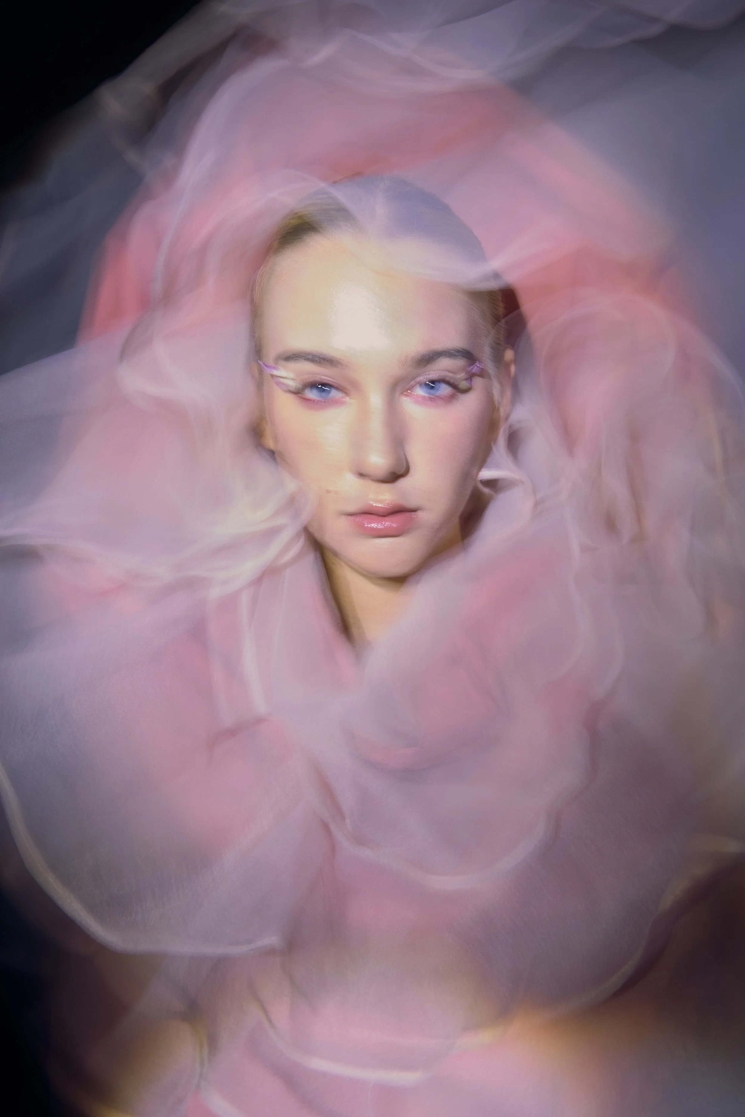 A woman with blonde hair and blue eyes surrounded by pink and purple flowing fabric or smoke, looking directly at the camera.