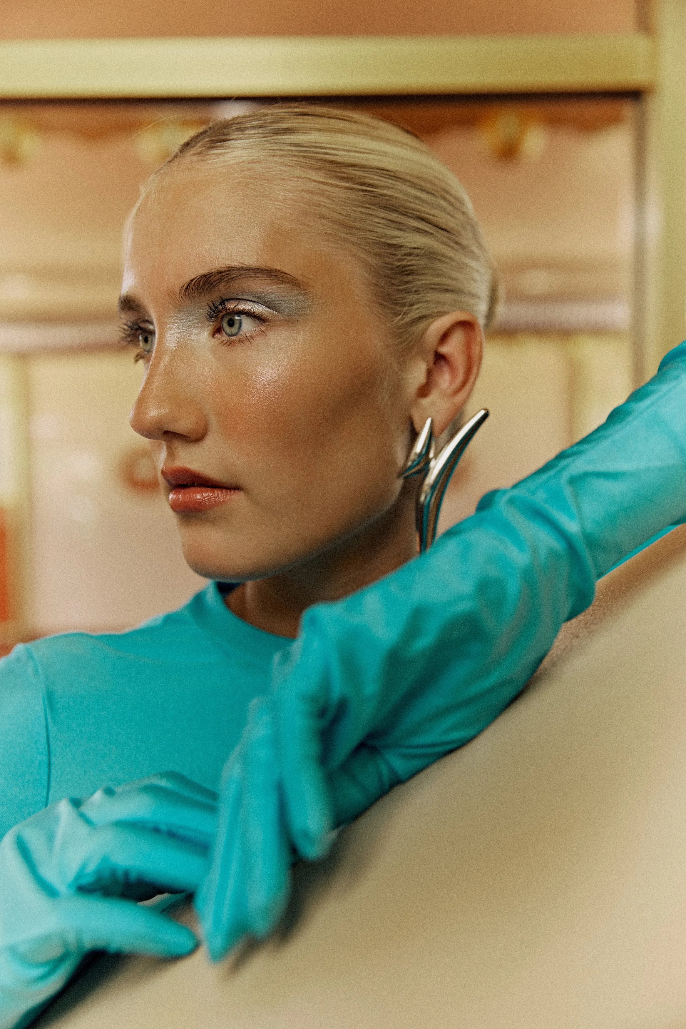 A woman with short blonde hair and light makeup, wearing a teal medical gown, sitting on a beige couch in a room with warm lighting.