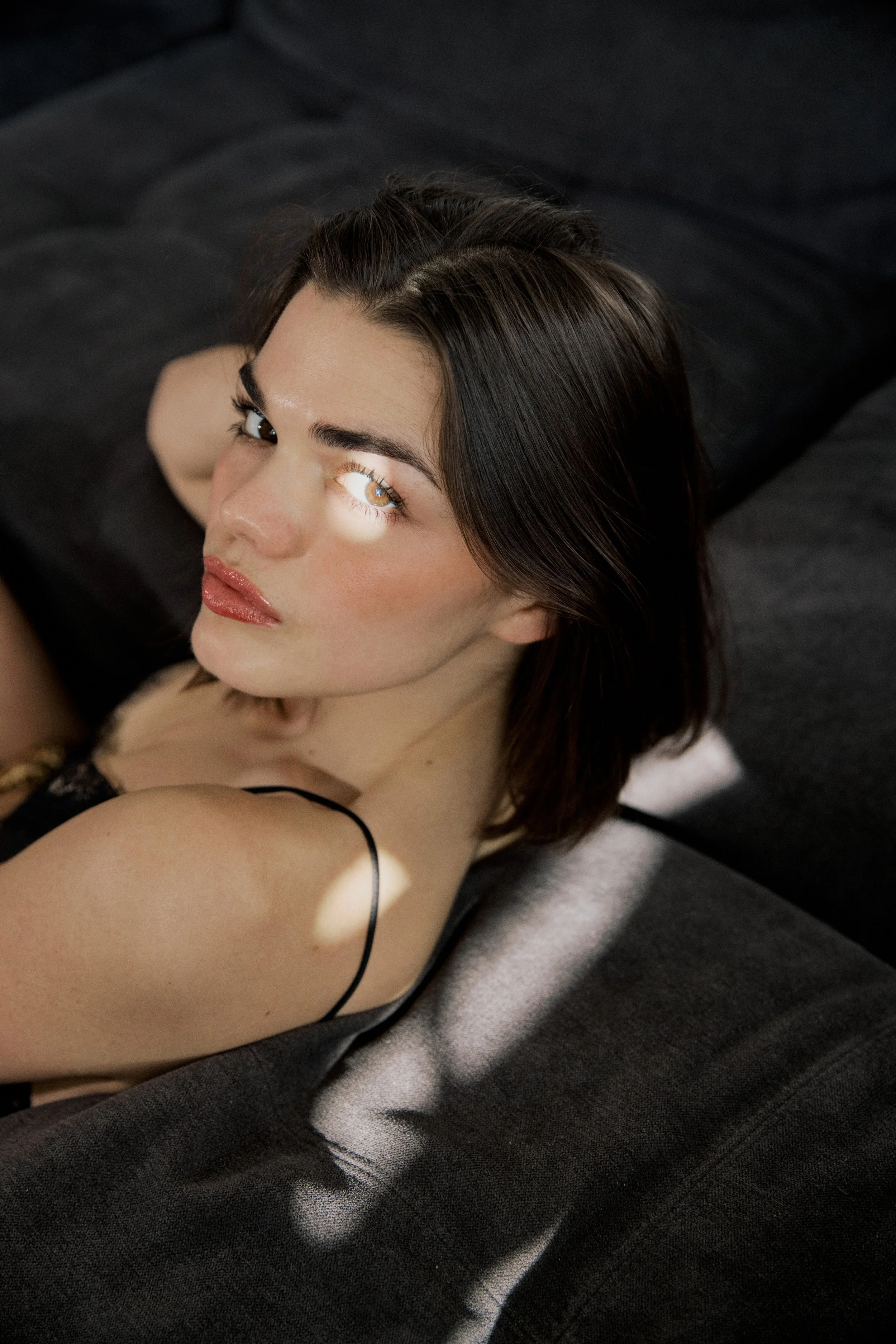 A woman with dark brown hair and light makeup lying on a black couch, looking up at the camera.