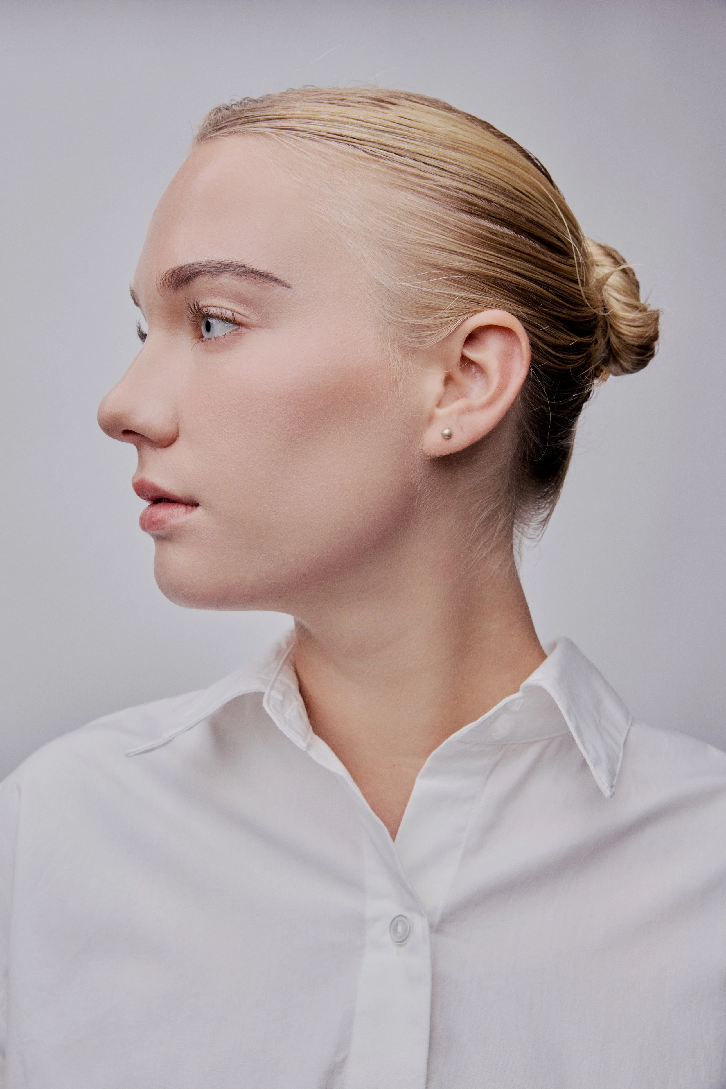 Profile of a woman with blonde hair tied in a bun, wearing a white collared shirt and small pearl earring, against a gray background.