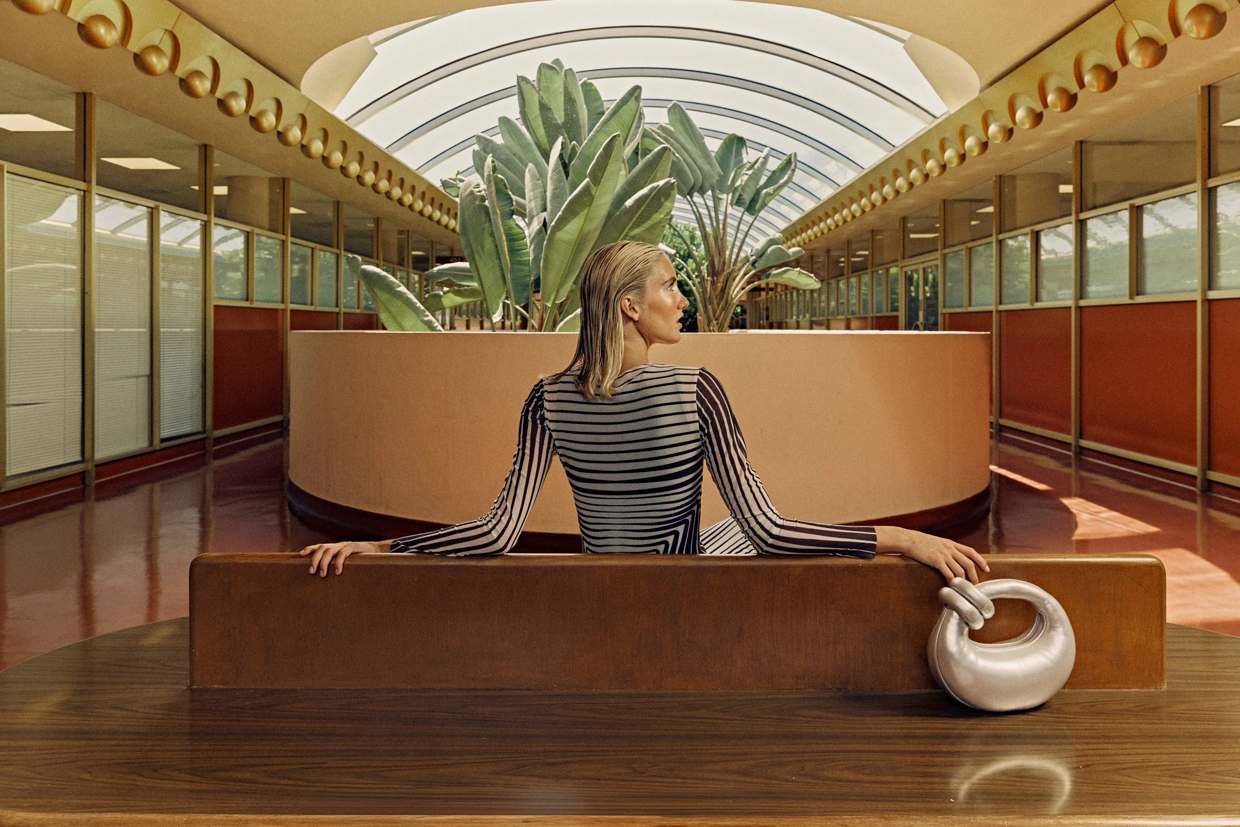 A woman with blonde hair in a striped dress is seated on a bench with her back straight, in an indoor space with a large green plant and a curved glass ceiling.