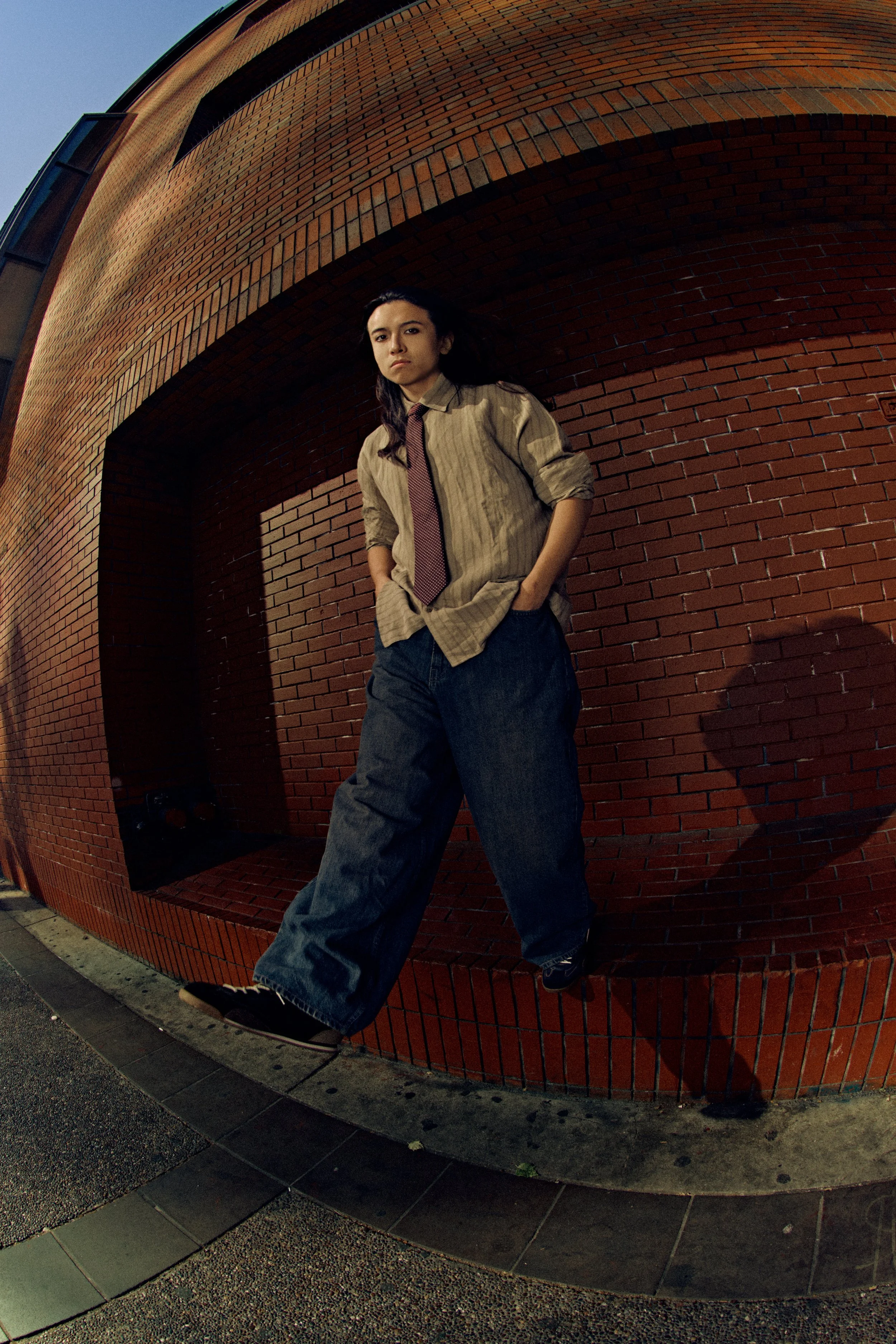 A young woman with dark hair in a loose ponytail, wearing a beige shirt, red tie, baggy blue jeans, and sneakers, leaning against a red brick wall on a sidewalk, with a building and a clear sky in the background.