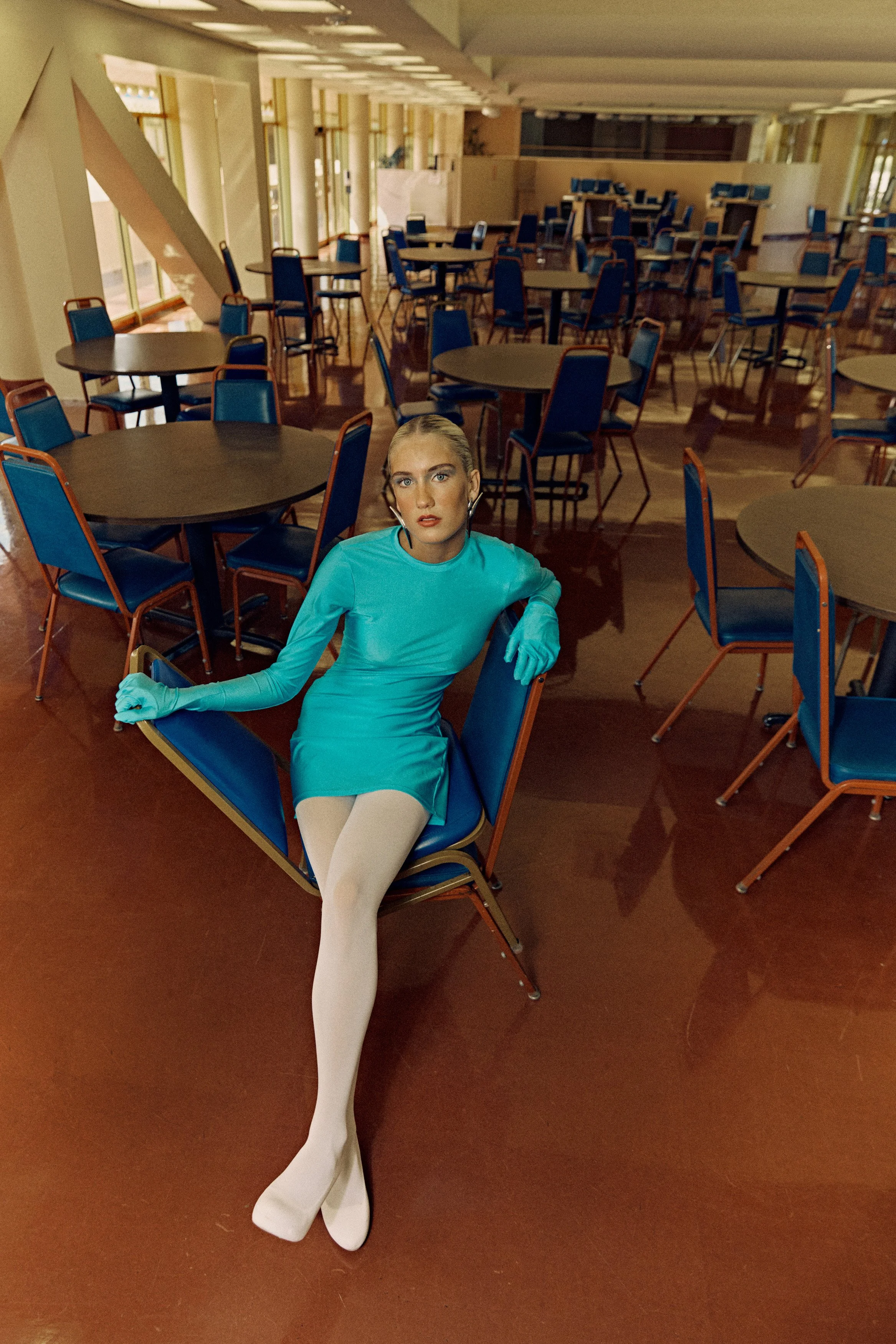 An empty spacious room with round tables and blue chairs, a woman with blonde hair in a teal dress and white tights sitting on a chair, with a serious expression.
