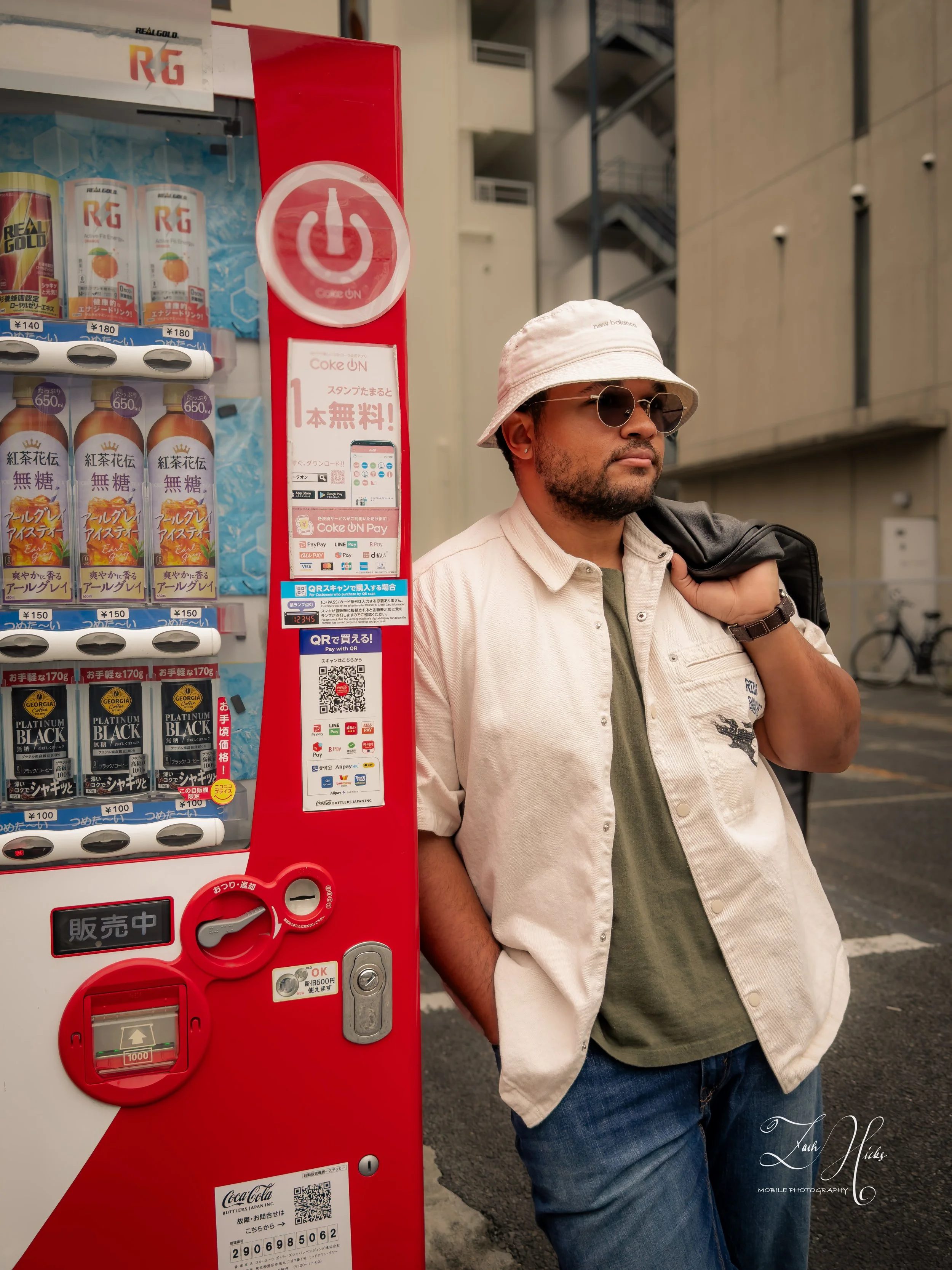 A man wearing sunglasses, a white bucket hat, an unbuttoned beige shirt over a green shirt, and jeans stands next to a red vending machine on a street. He is holding a black bag over his shoulder.