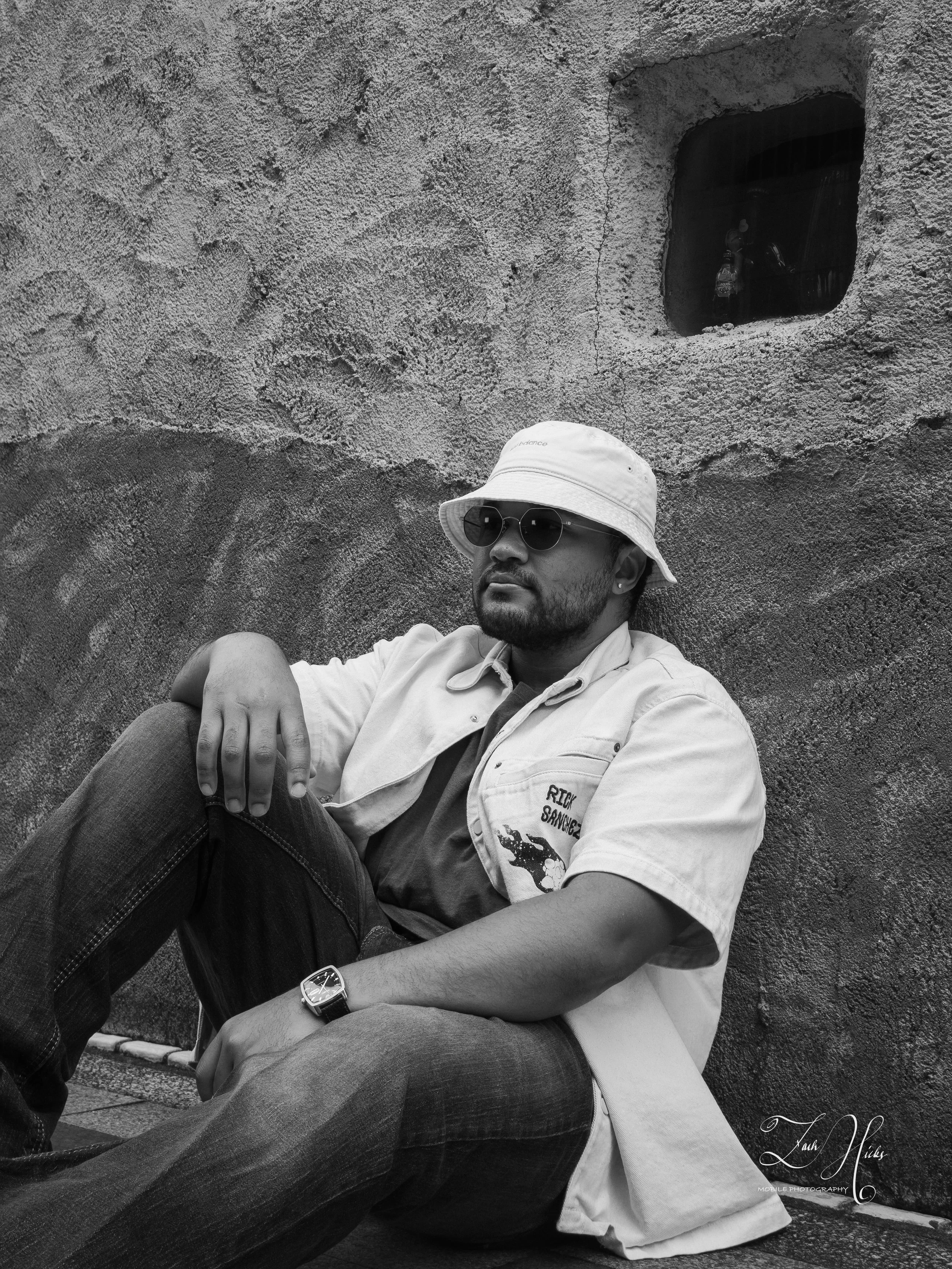 A man wearing sunglasses, a bucket hat, a short sleeve jacket, and jeans, sitting against a textured wall with a small window in the upper right corner.