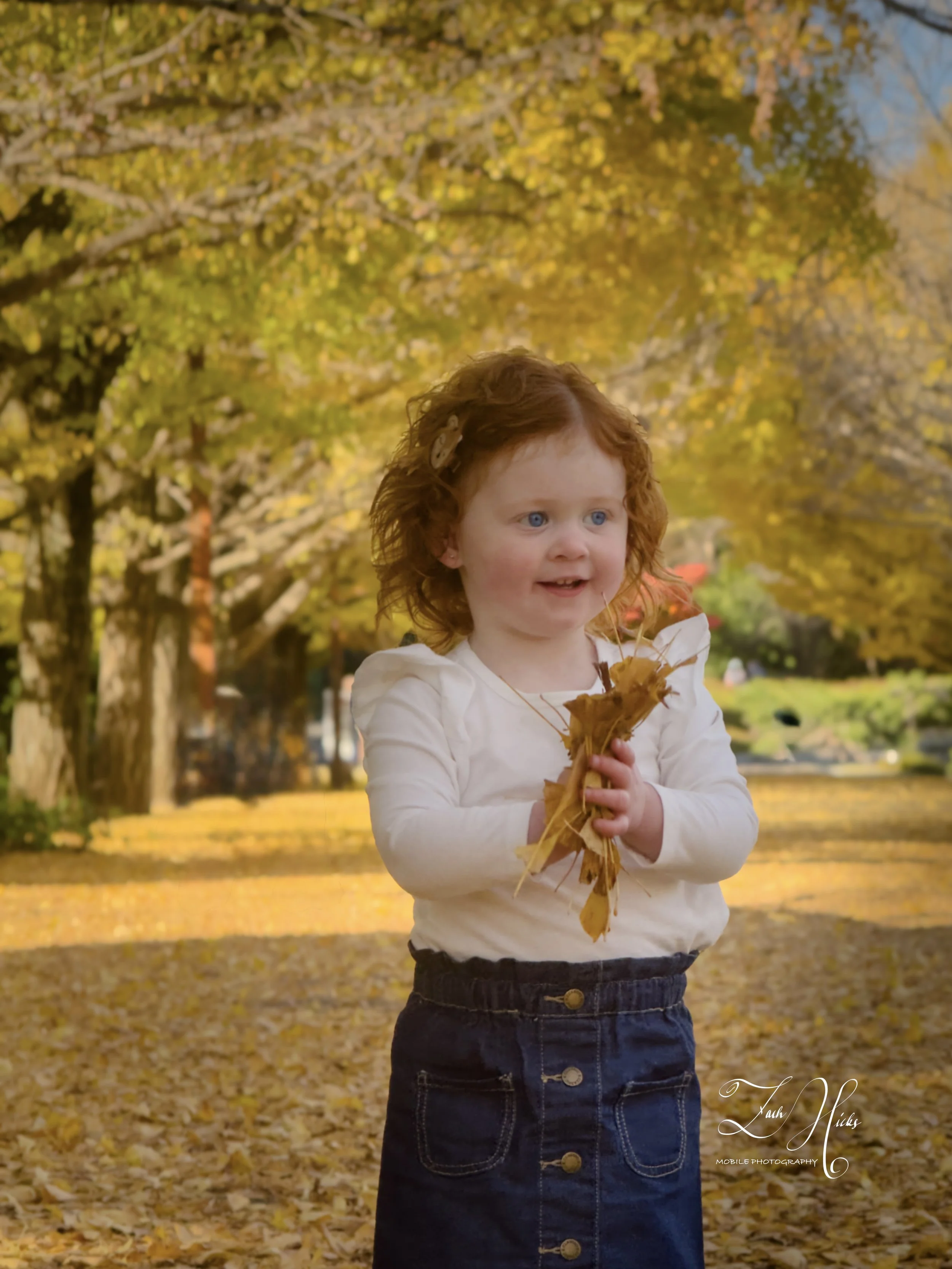 A young girl with red hair and blue eyes holding a large leaf in a park with autumn trees and fallen leaves.