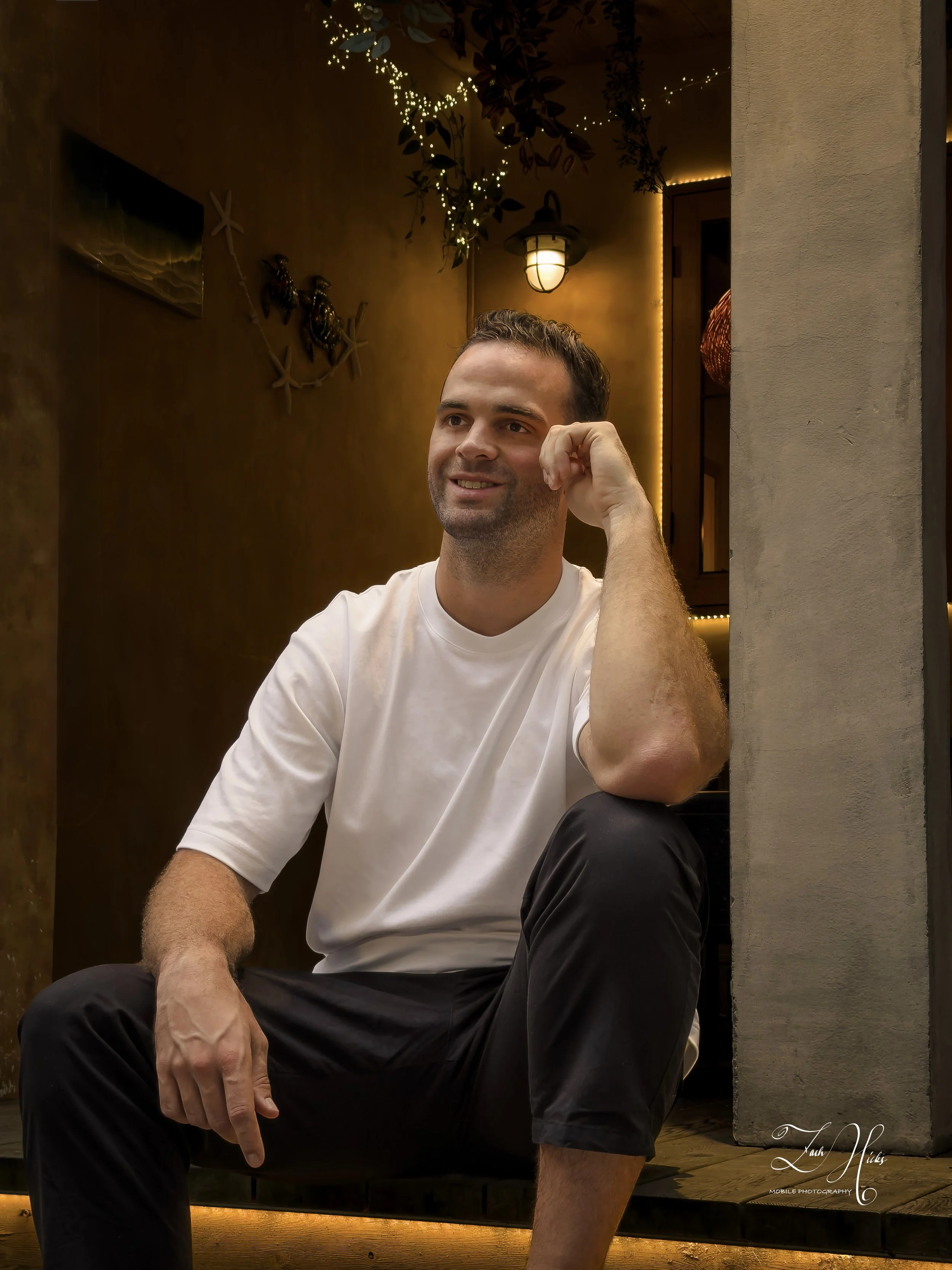 A man sitting in a cozy, warmly lit space, wearing a white t-shirt and black pants, smiling thoughtfully with his left hand resting on his face.