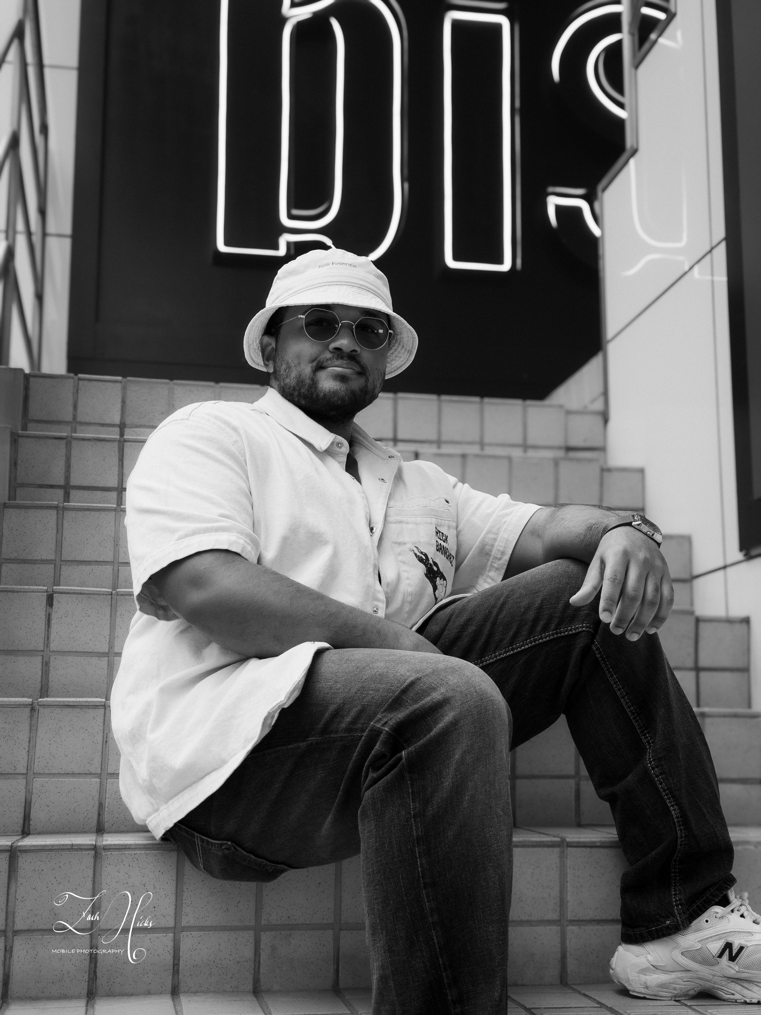 A man sitting on tiled steps in front of a neon sign with the word 'DIE' in large letters. He is wearing sunglasses, a white bucket hat, a short-sleeved shirt, jeans, and sneakers.