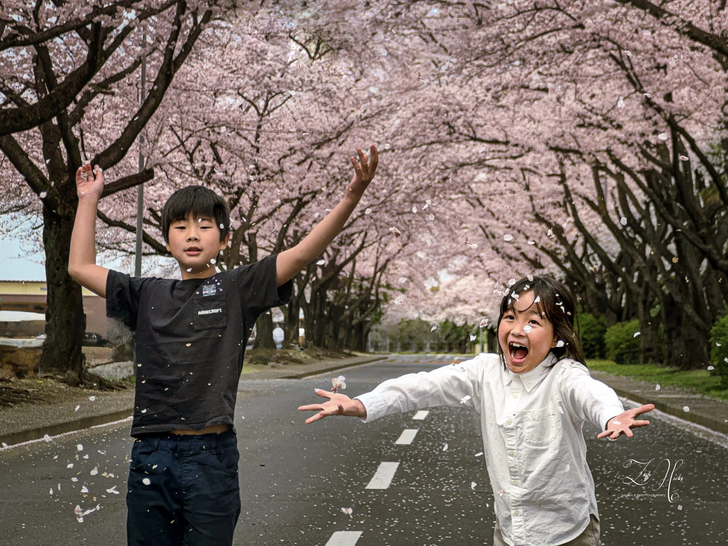 Two children, a boy and a girl, playing in the street with cherry blossom petals falling around them during daytime.
