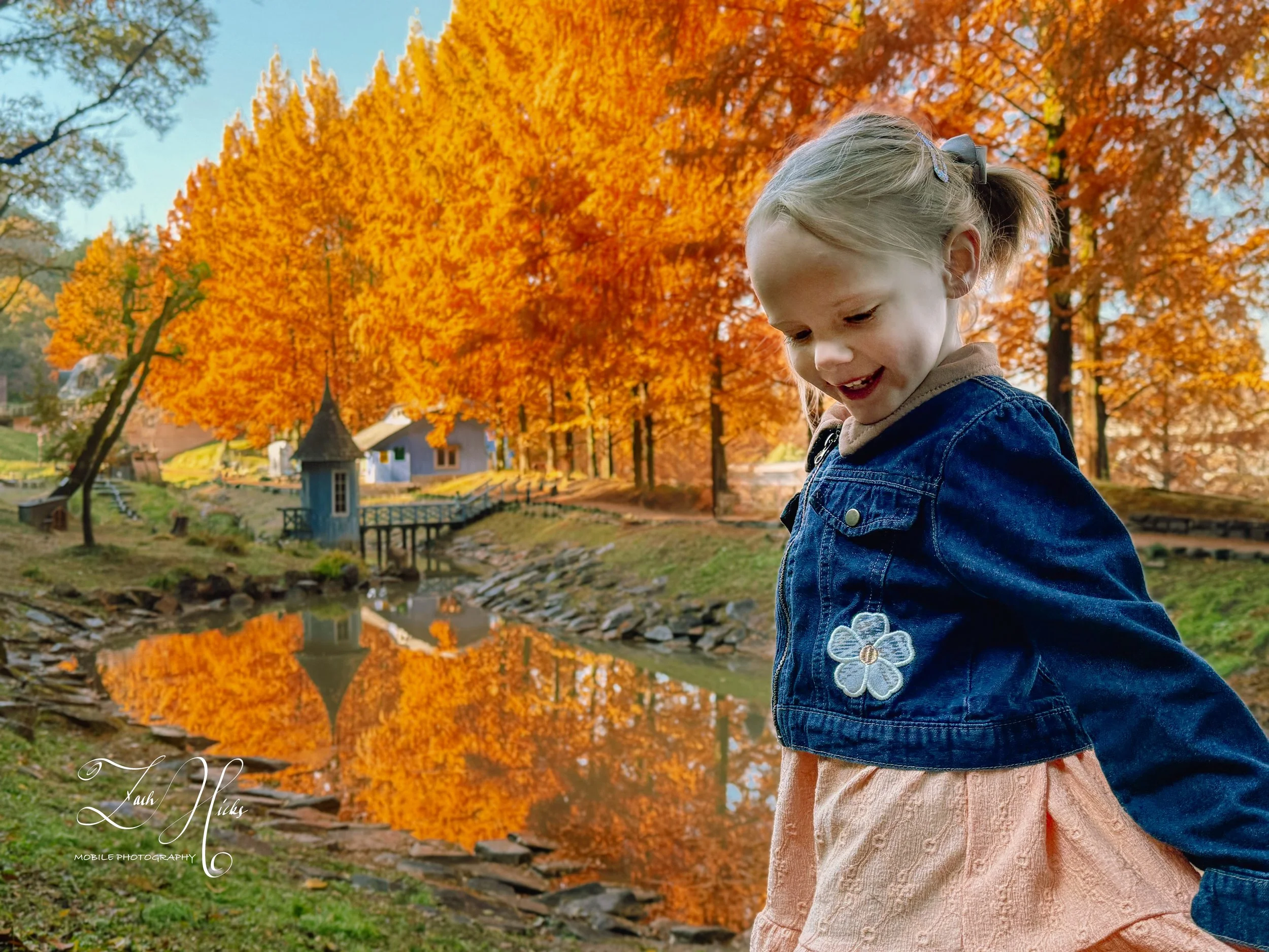 A young girl smiling and looking down, wearing a denim jacket with an embroidered flower, standing near a pond with autumn orange trees in the background.