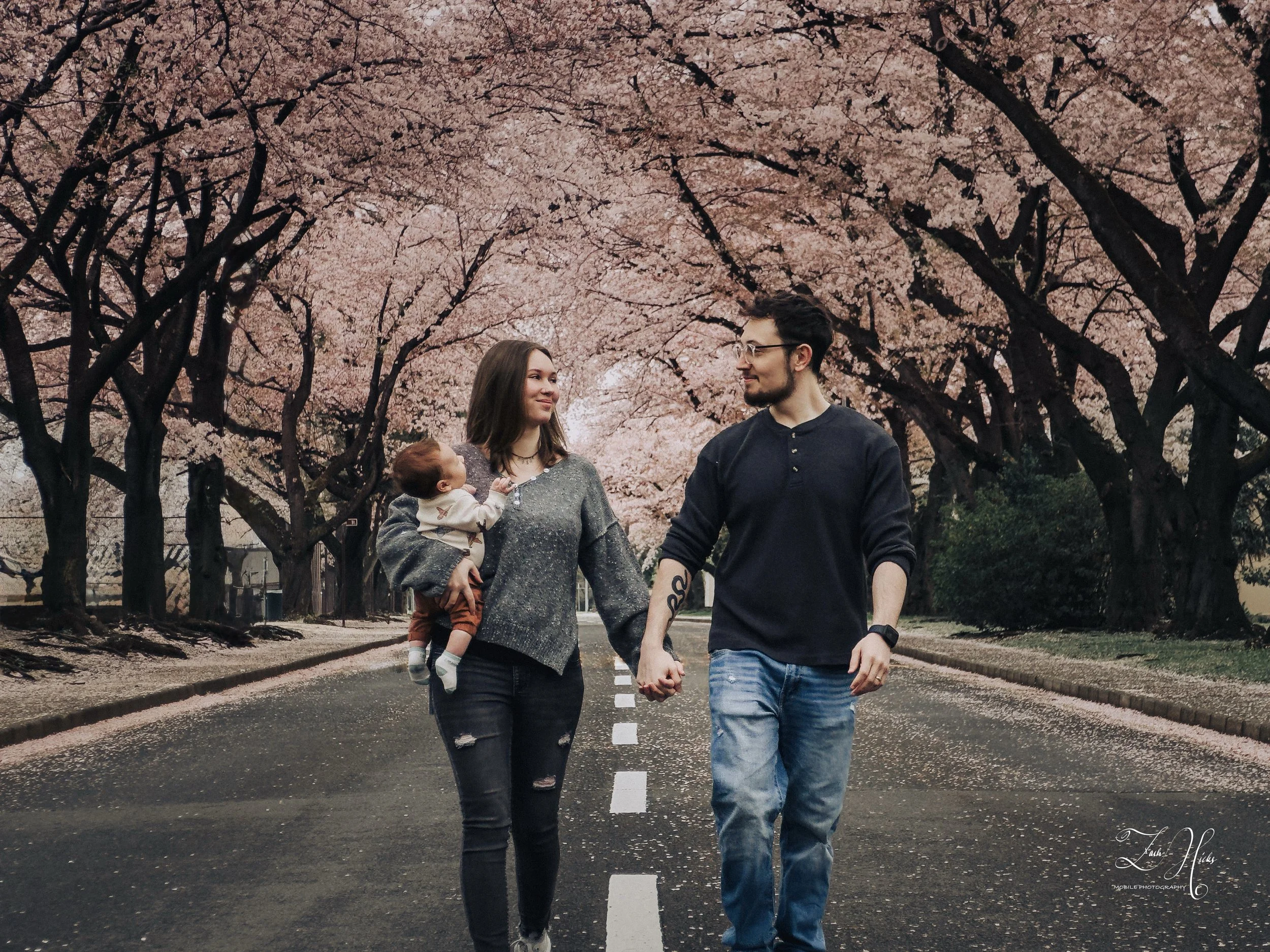 A young family walking hand in hand on a tree-lined street during cherry blossom season. The mother holds a baby, and they all enjoy the scenic surroundings.