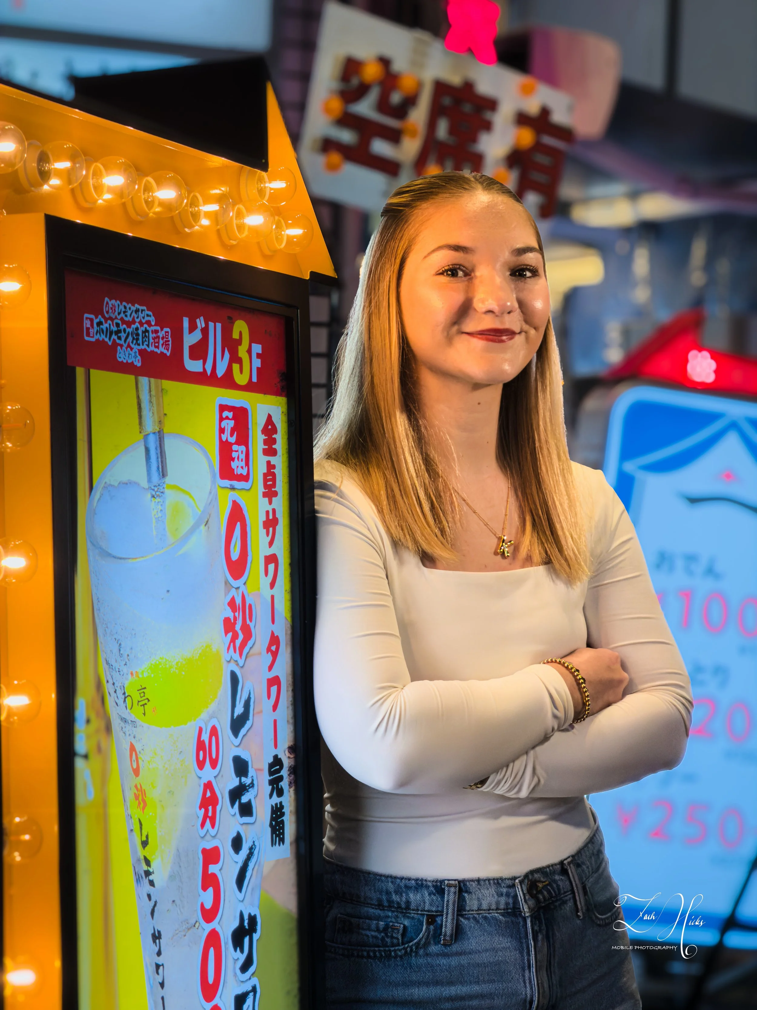 A young woman with long blonde hair standing next to a brightly lit, colorful advertisement board at what appears to be a street food or night market. She is smiling, arms crossed, wearing a cream-colored top and blue jeans, with her hair neatly tuck