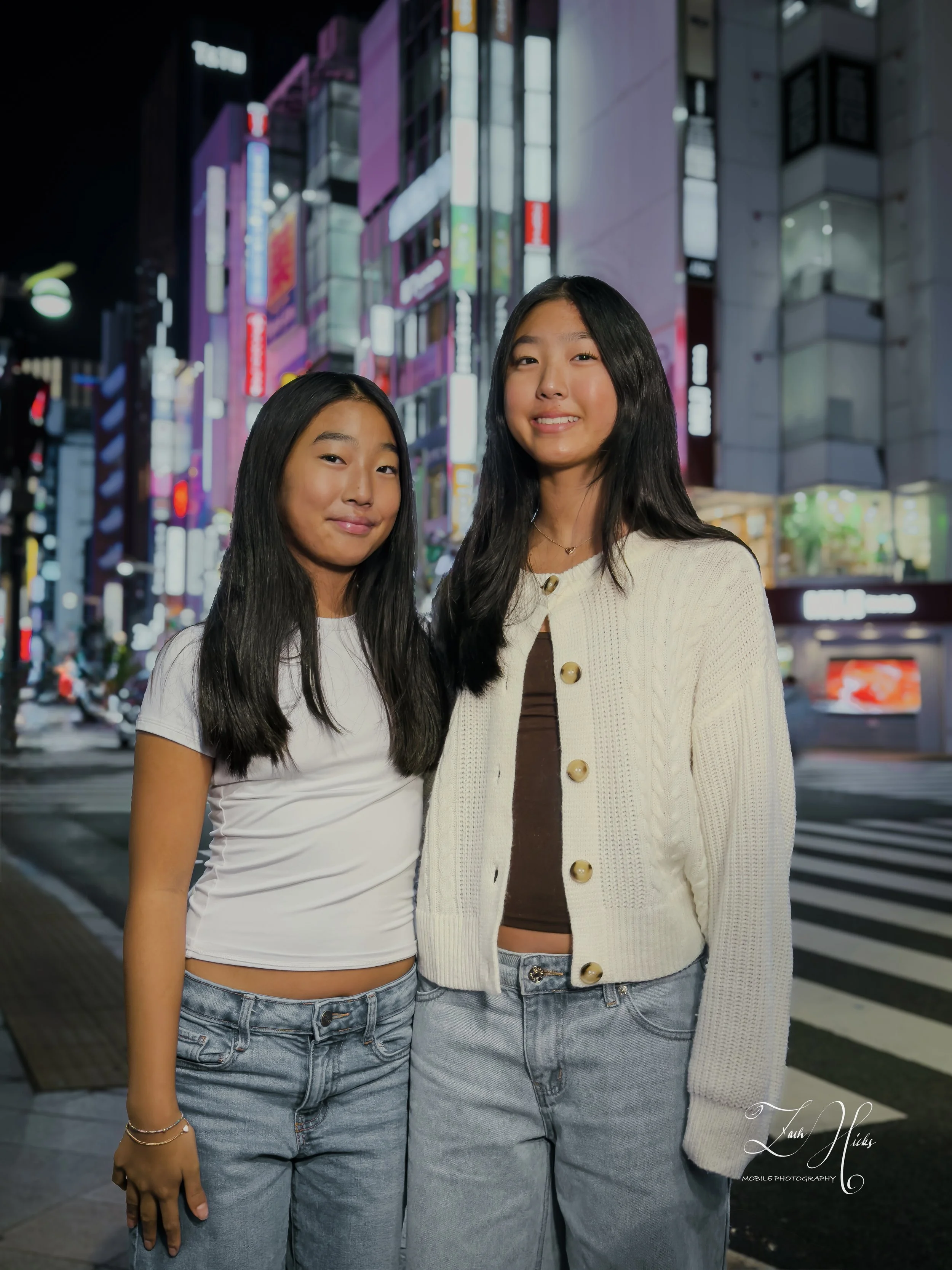 Two young women standing on a city street at night with brightly lit buildings and signs in the background, smiling at the camera.