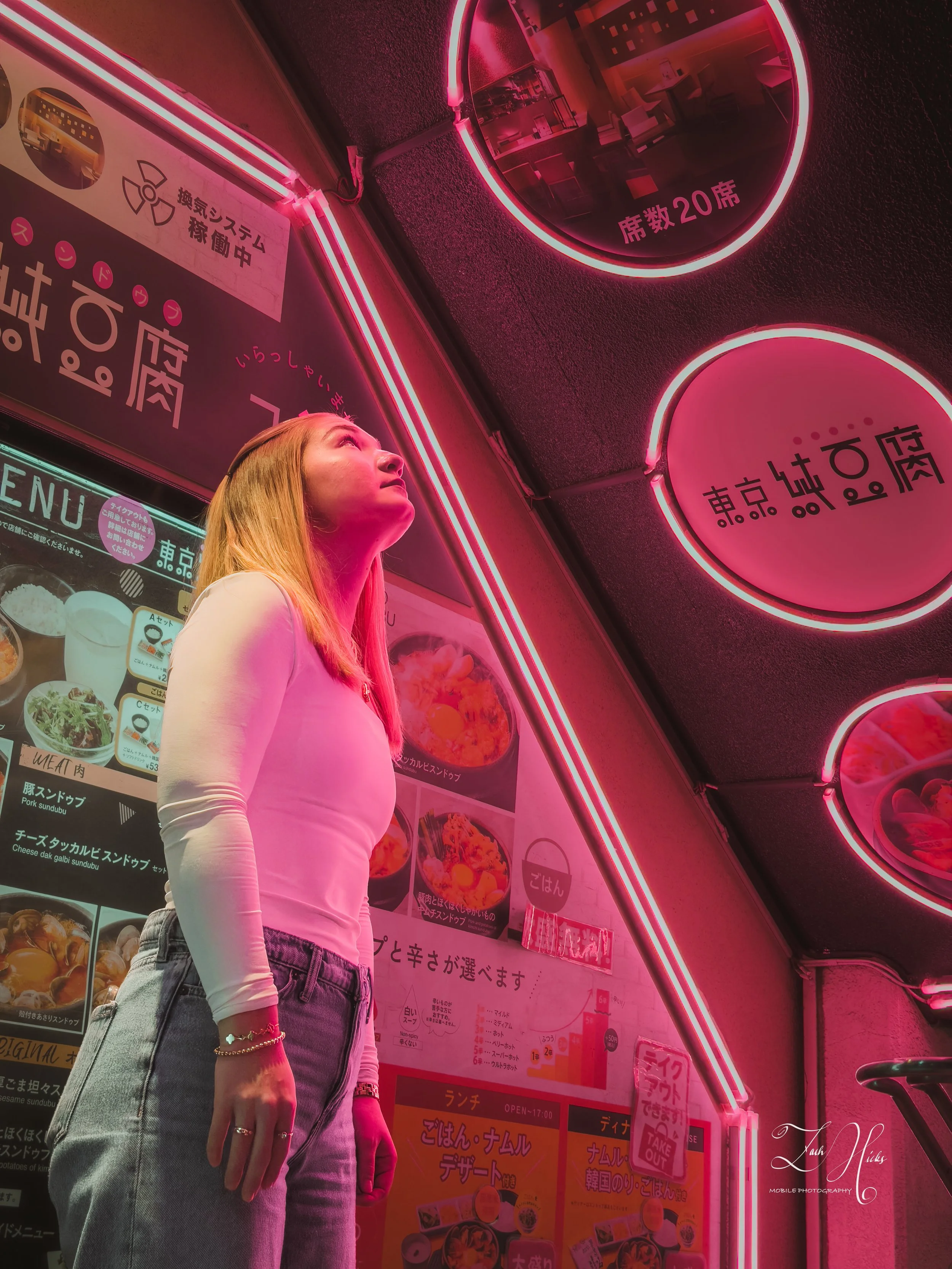 A young woman with blonde hair, wearing a white long-sleeve top and light blue jeans, stands in front of Japanese restaurant signs illuminated with pink neon lights.
