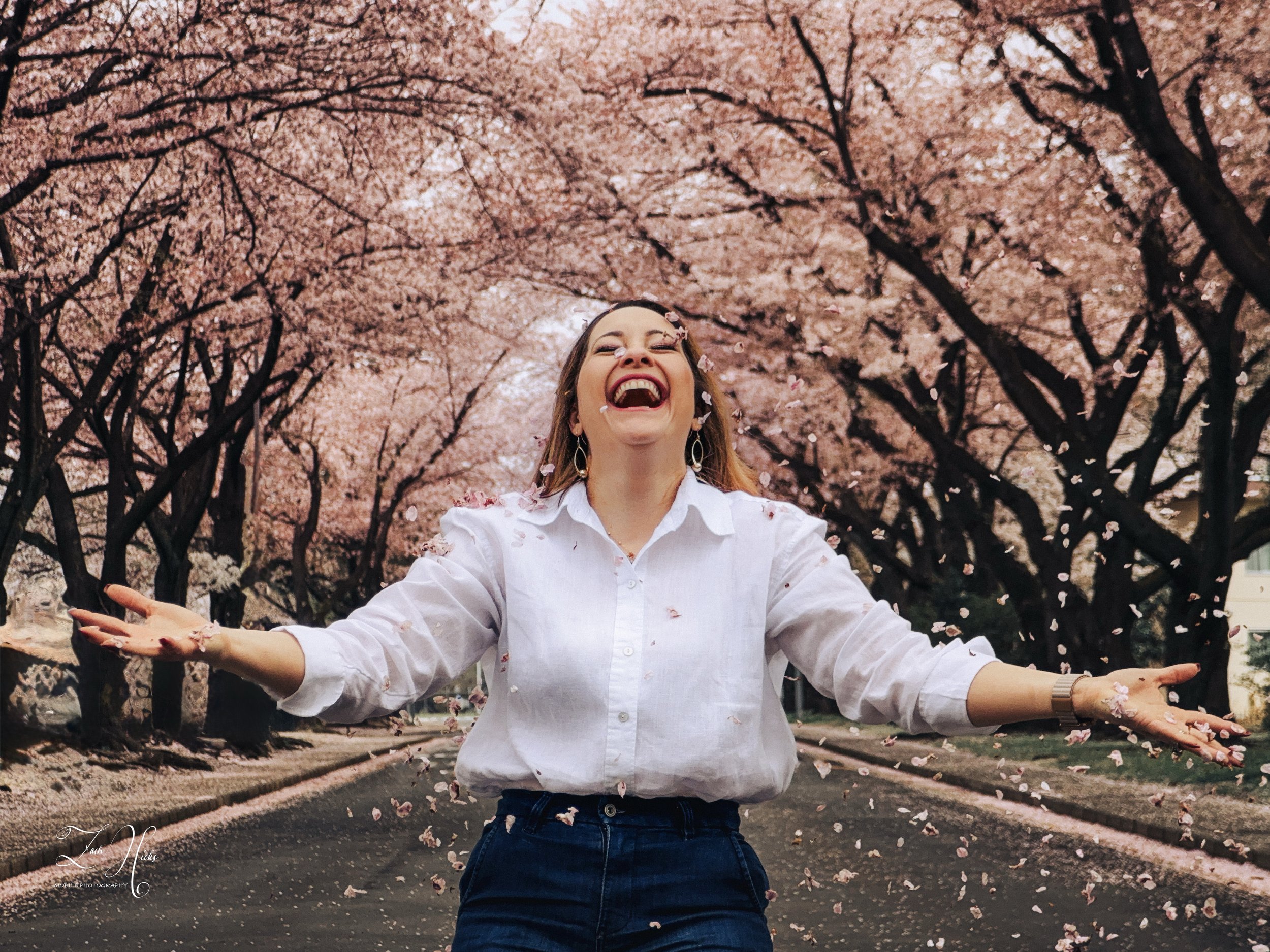 A woman with arms outstretched, smiling, standing under cherry blossom trees with pink flowers, petals falling around her.