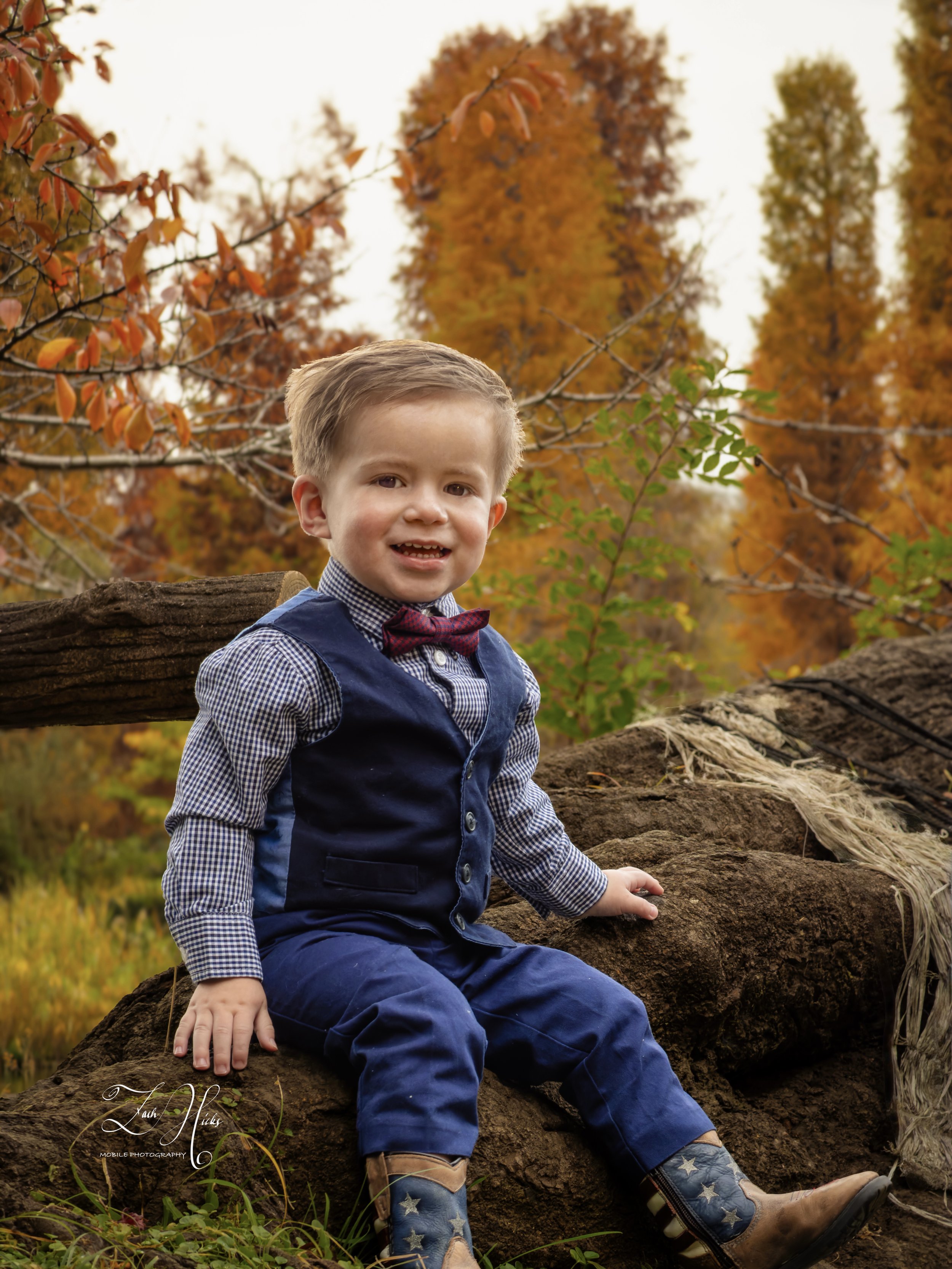 Young boy sitting outdoors on a log during fall, wearing a checkered shirt, navy vest, red bow tie, blue pants, and cowboy boots. Autumn trees with orange and yellow leaves in the background.