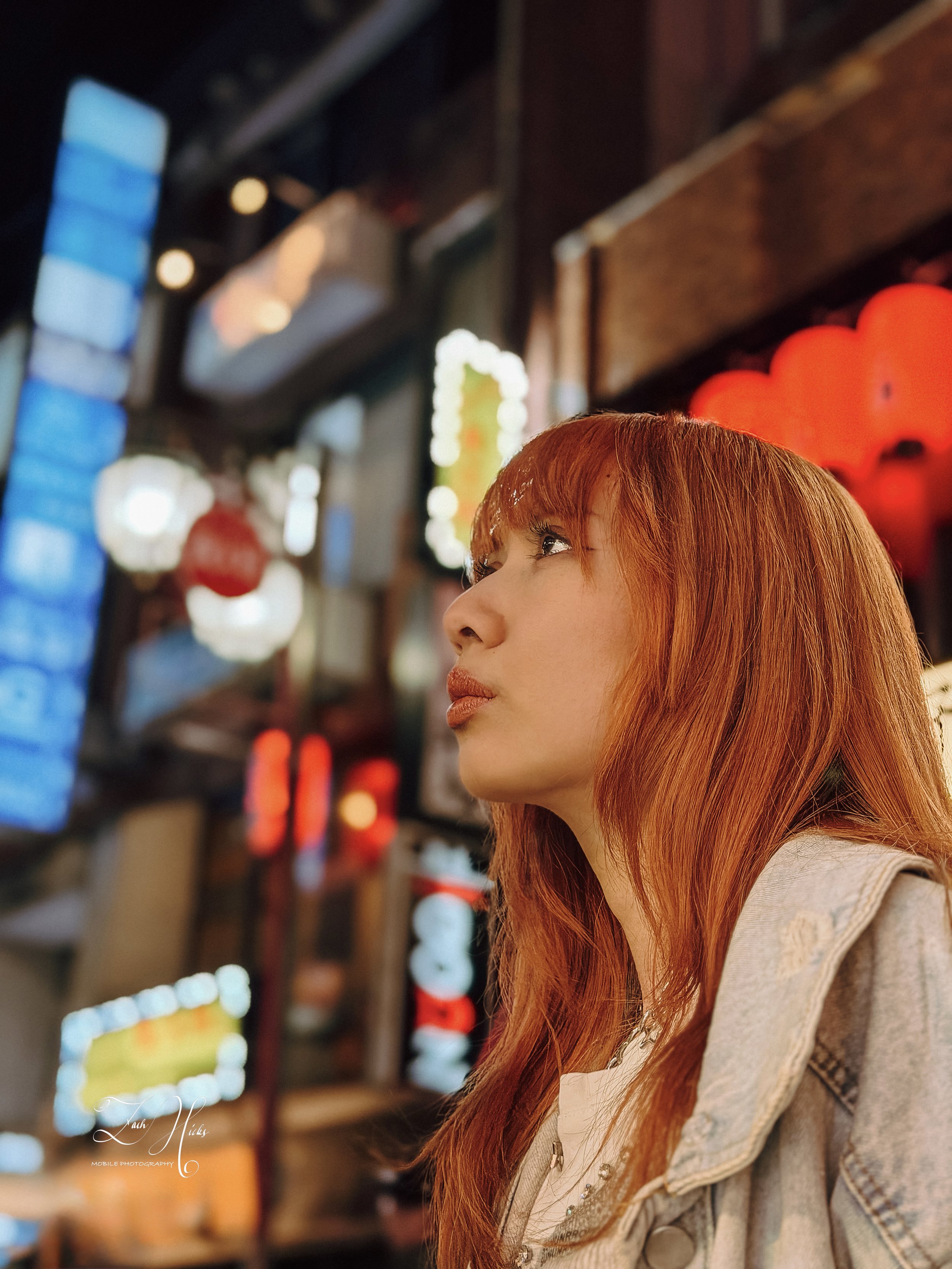 A woman with long red hair is looking to her left in a brightly lit, colorful setting with neon and decorative lights.