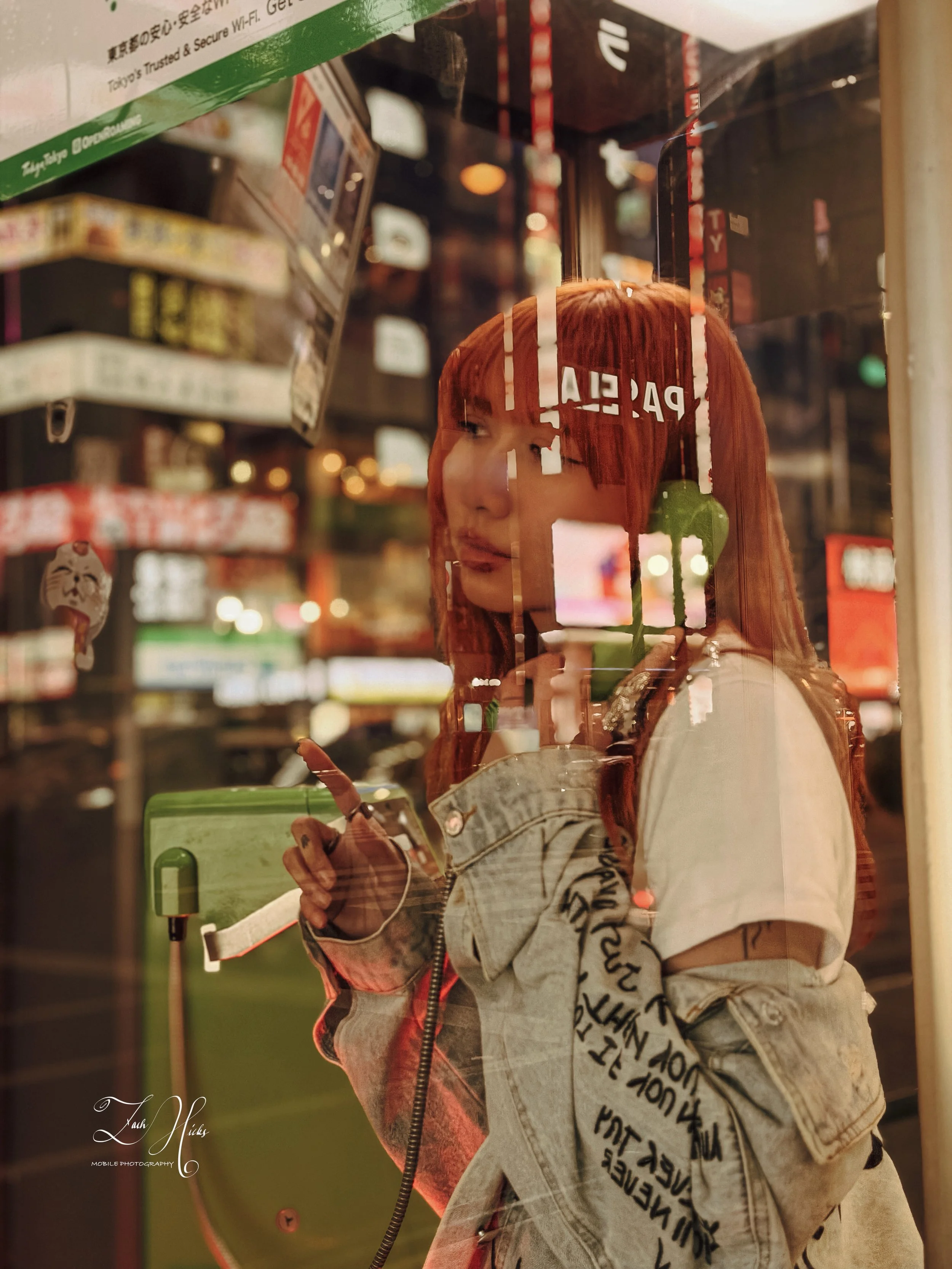 A young woman with long red hair and tattoos, dressed in a beige jacket, looks through a glass window at night. Reflected city lights and neon signs create a colorful, layered effect on the glass.