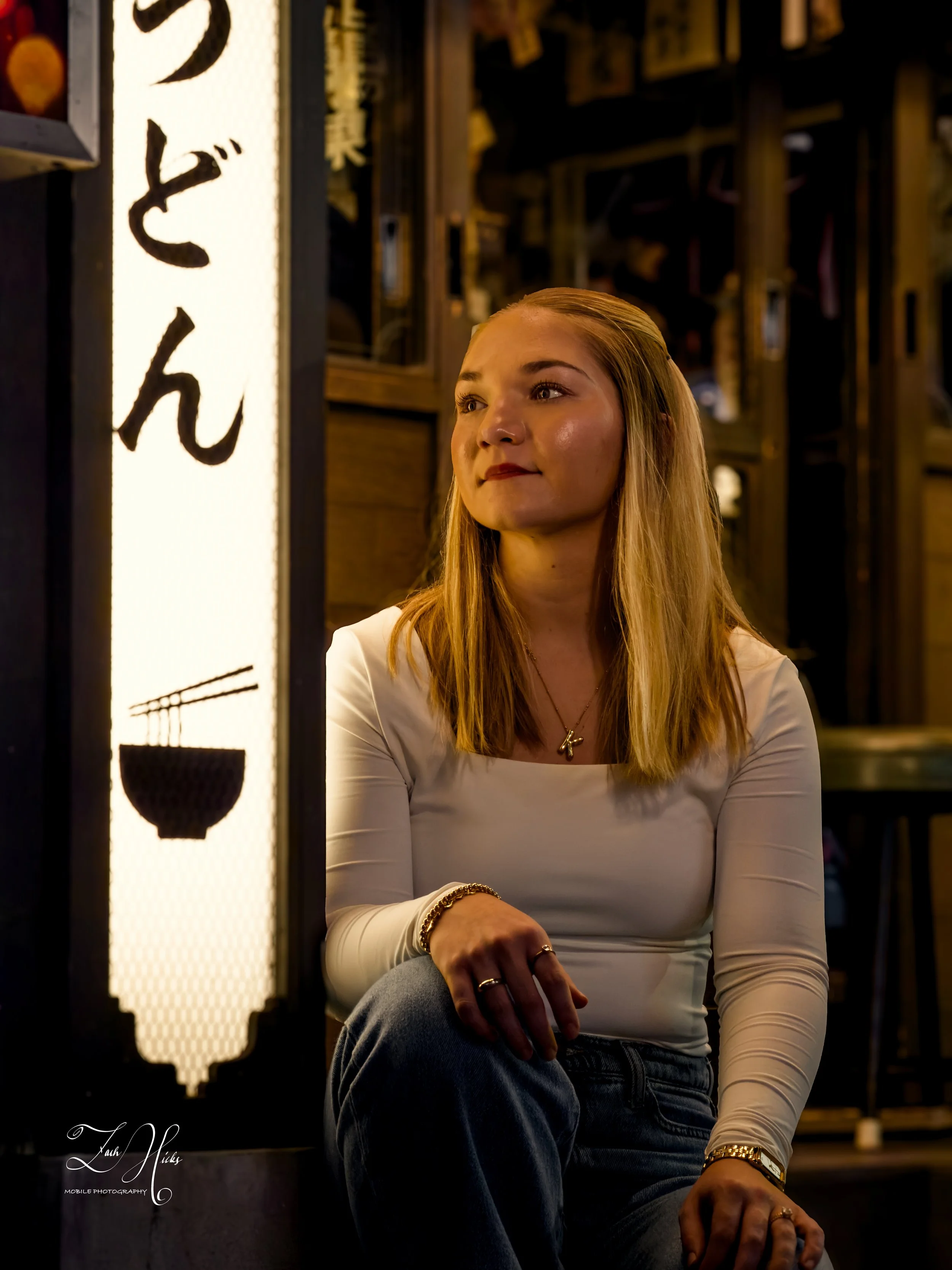 A young woman with long blonde hair sitting in a restaurant or bar with wooden decor. She is wearing a white long sleeve top and jeans, with jewelry including rings, a bracelet, and a necklace. There is a lit vertical sign with Japanese characters an