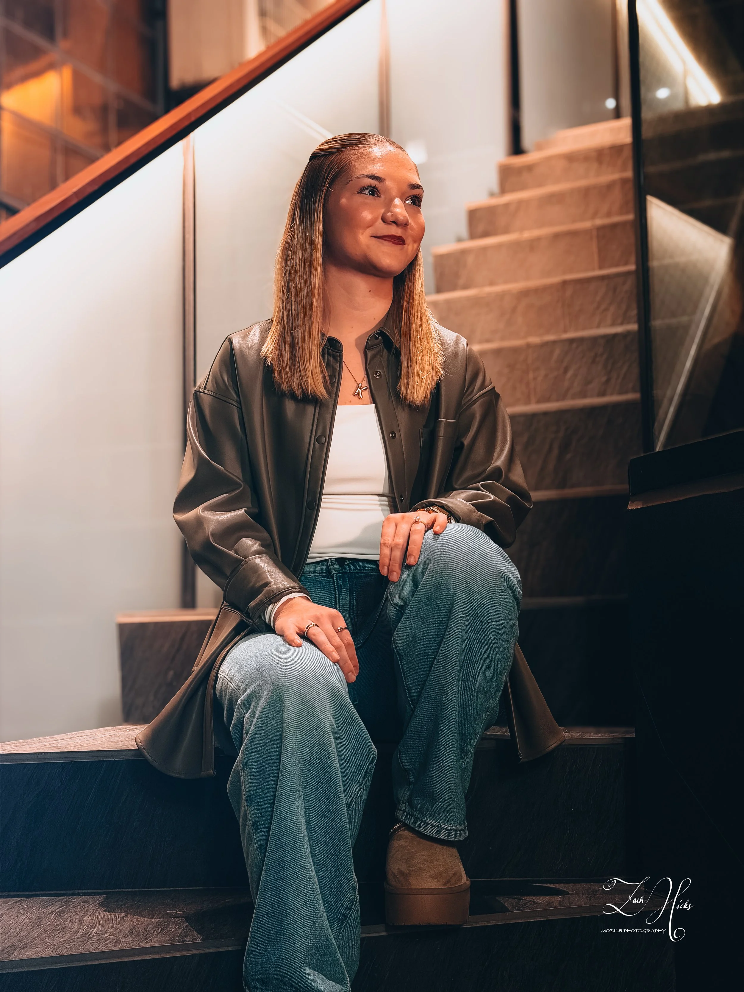 A woman with shoulder-length straight hair, wearing a leather jacket and jeans, sitting on stairs in a modern indoor setting with wooden and glass elements.