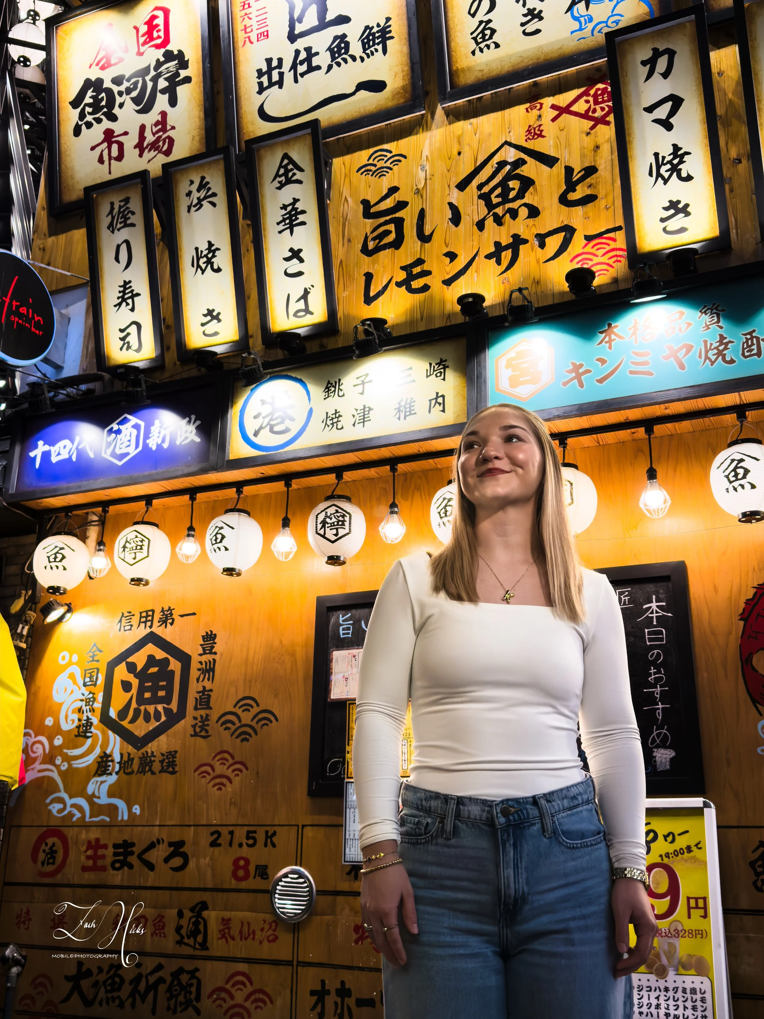 A woman in a white top and jeans standing in front of a Japanese restaurant with illuminated signs and lanterns.