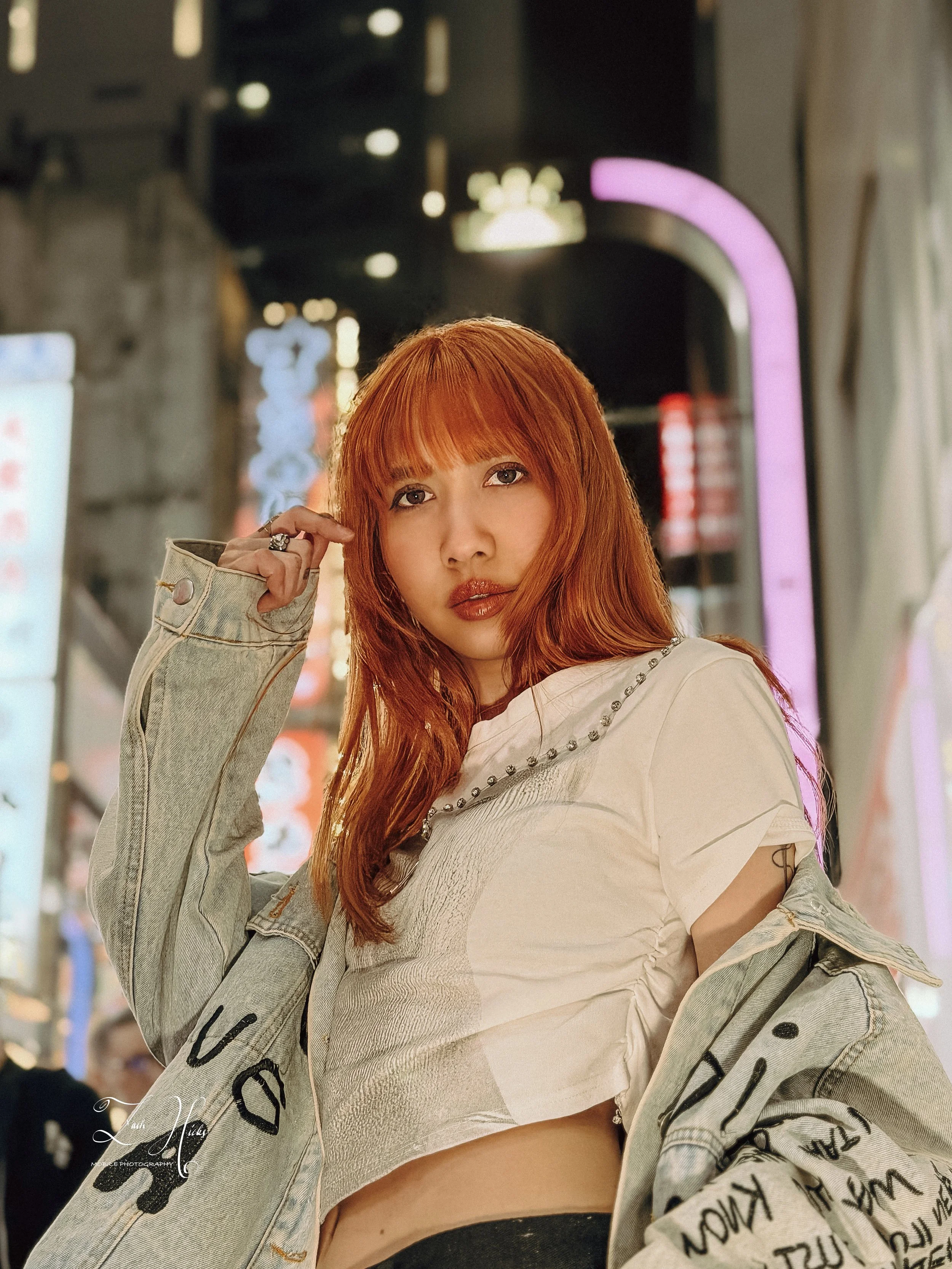 A young woman with red hair and fair skin in an urban setting at night, wearing a white rococo styled top and a distressed denim jacket with black handwritten text, standing in front of neon signs.