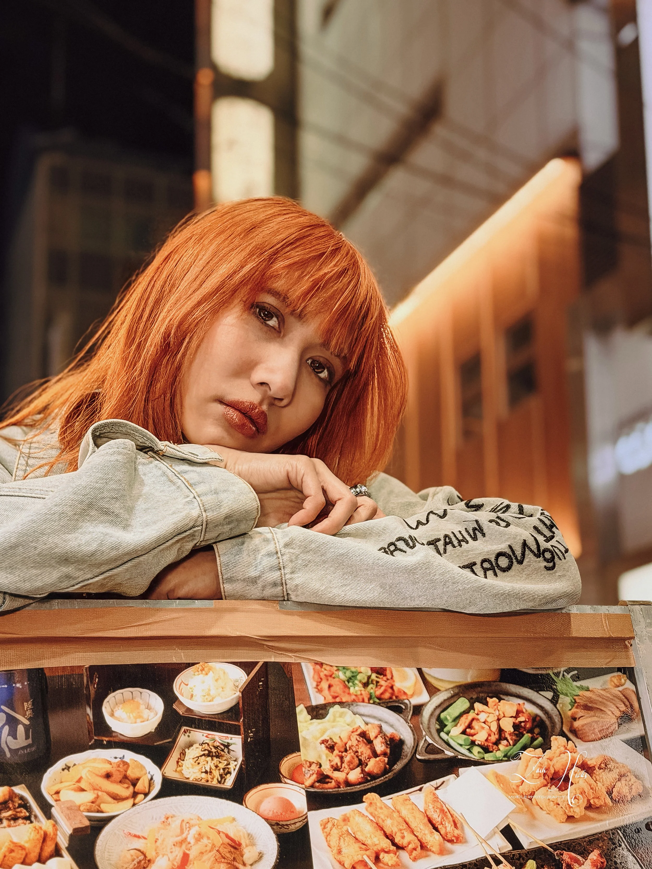 A red-haired woman resting her head on her crossed arms at a restaurant table with various Asian dishes.