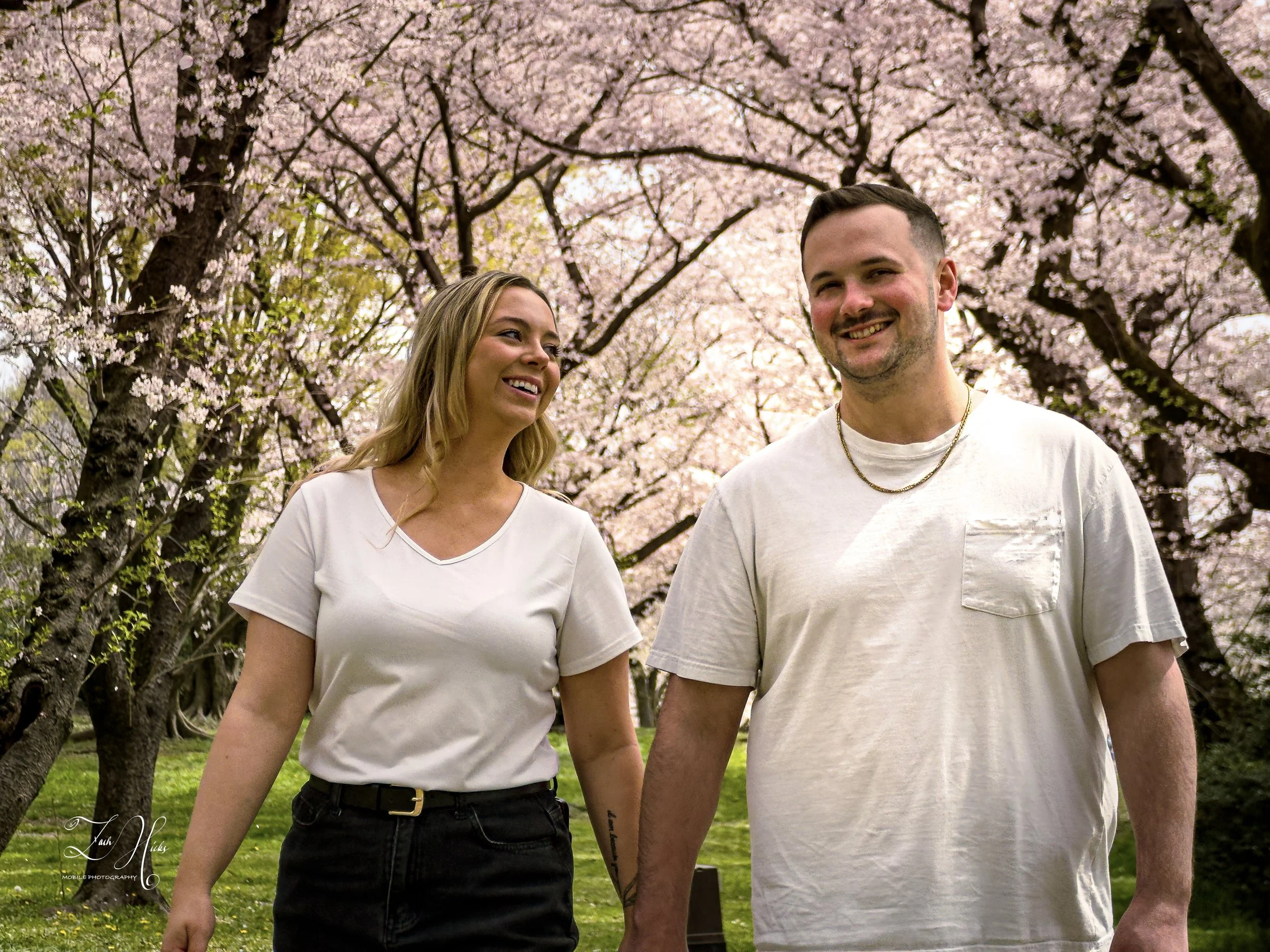 A smiling man and woman walking hand in hand through a park with blooming cherry blossom trees.