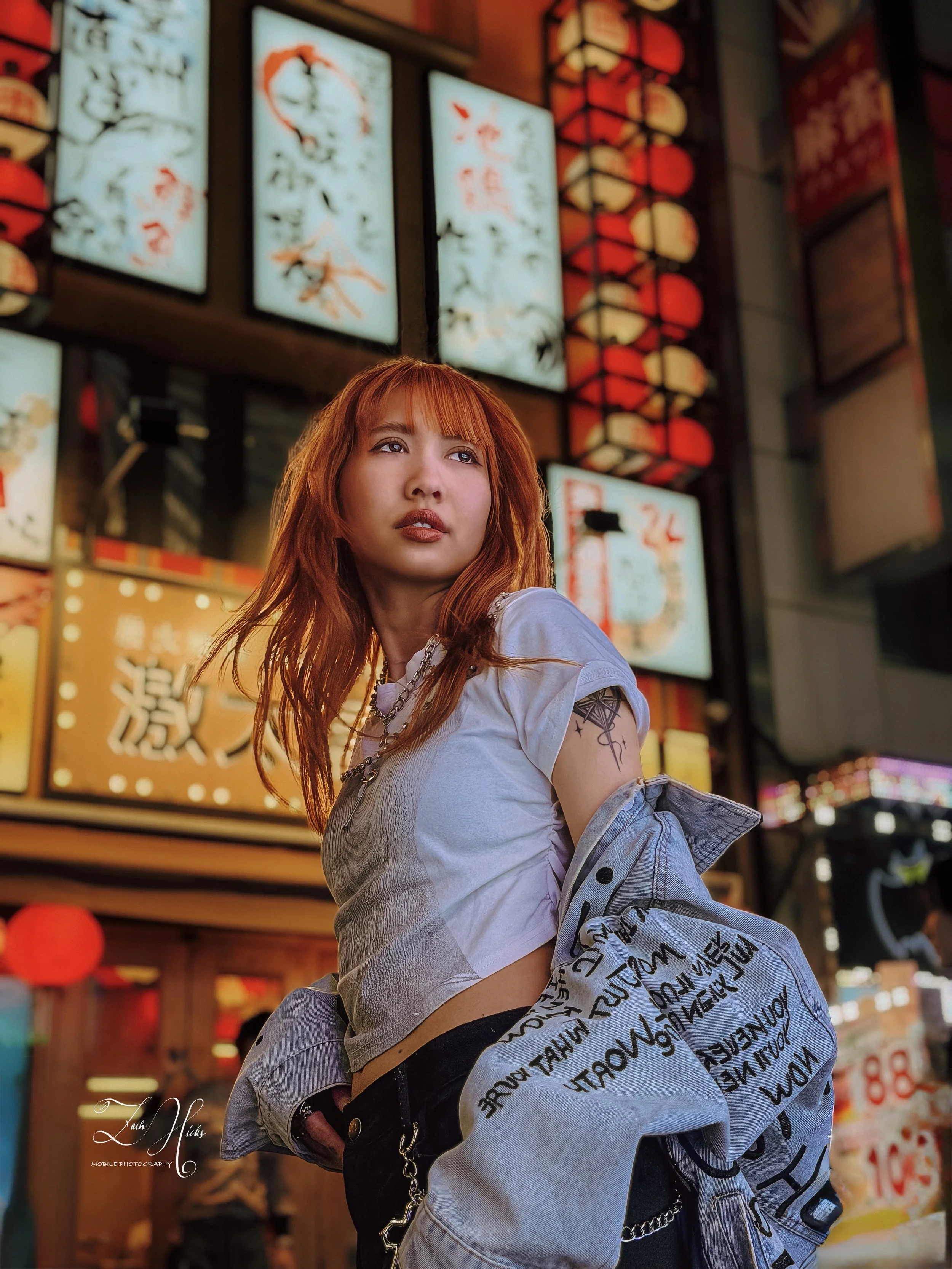 A young woman with red hair, tattoos, and wearing a white t-shirt and denim jacket, stands in front of brightly lit Japanese signage and lanterns in an urban area at night.