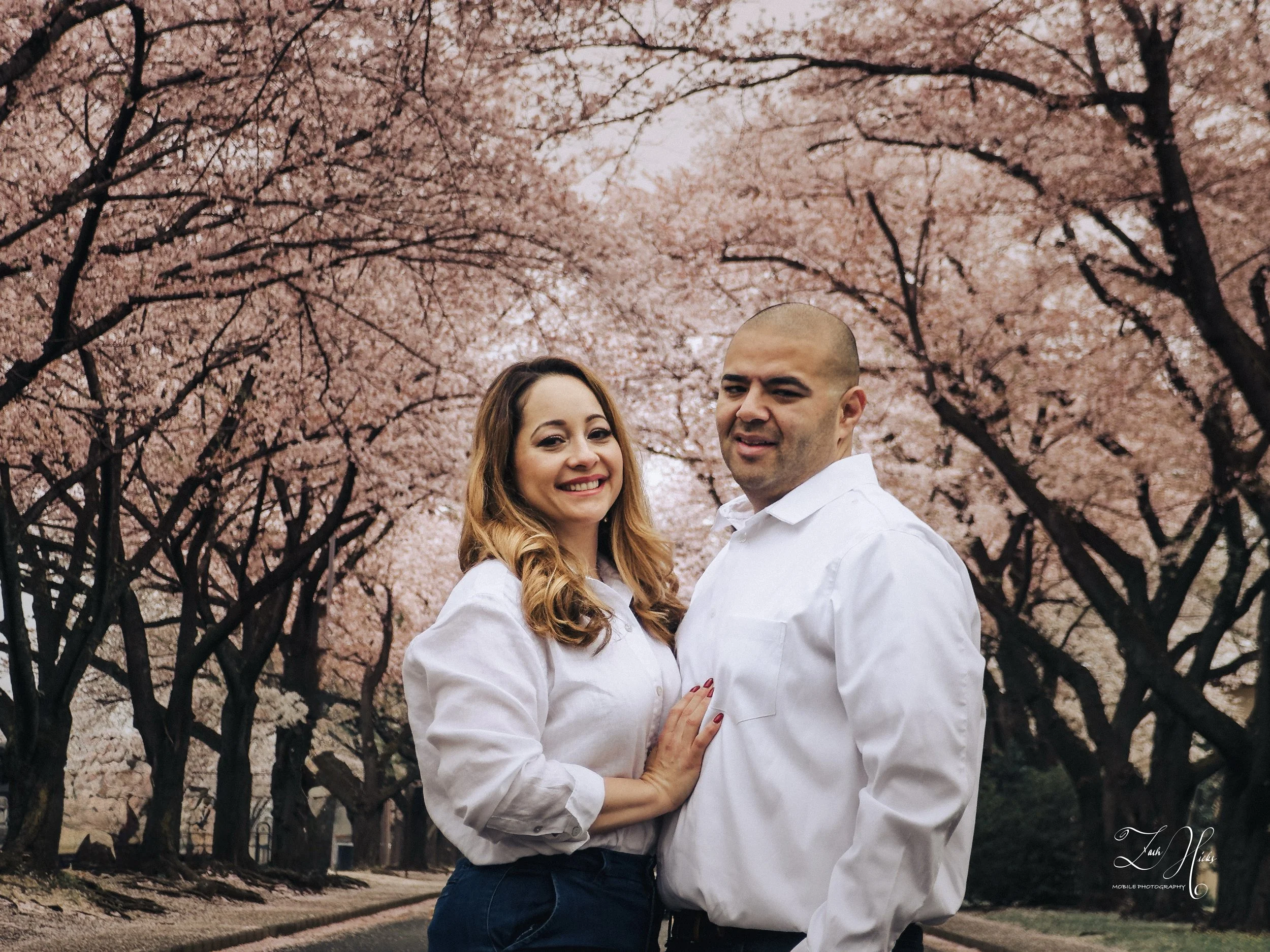 A smiling woman and a serious-looking man stand close together on a tree-lined path during spring, with pink cherry blossoms in full bloom overhead.