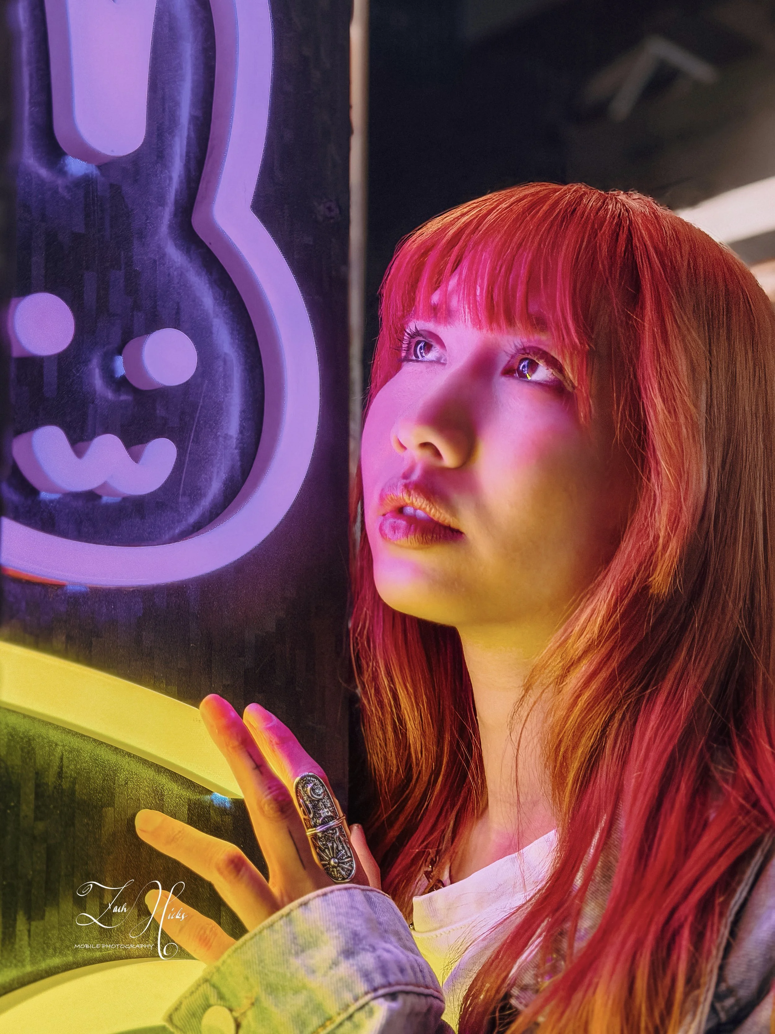 A young woman with red hair and bangs looking at neon signage in a dimly lit environment, with colorful lights reflecting on her face.