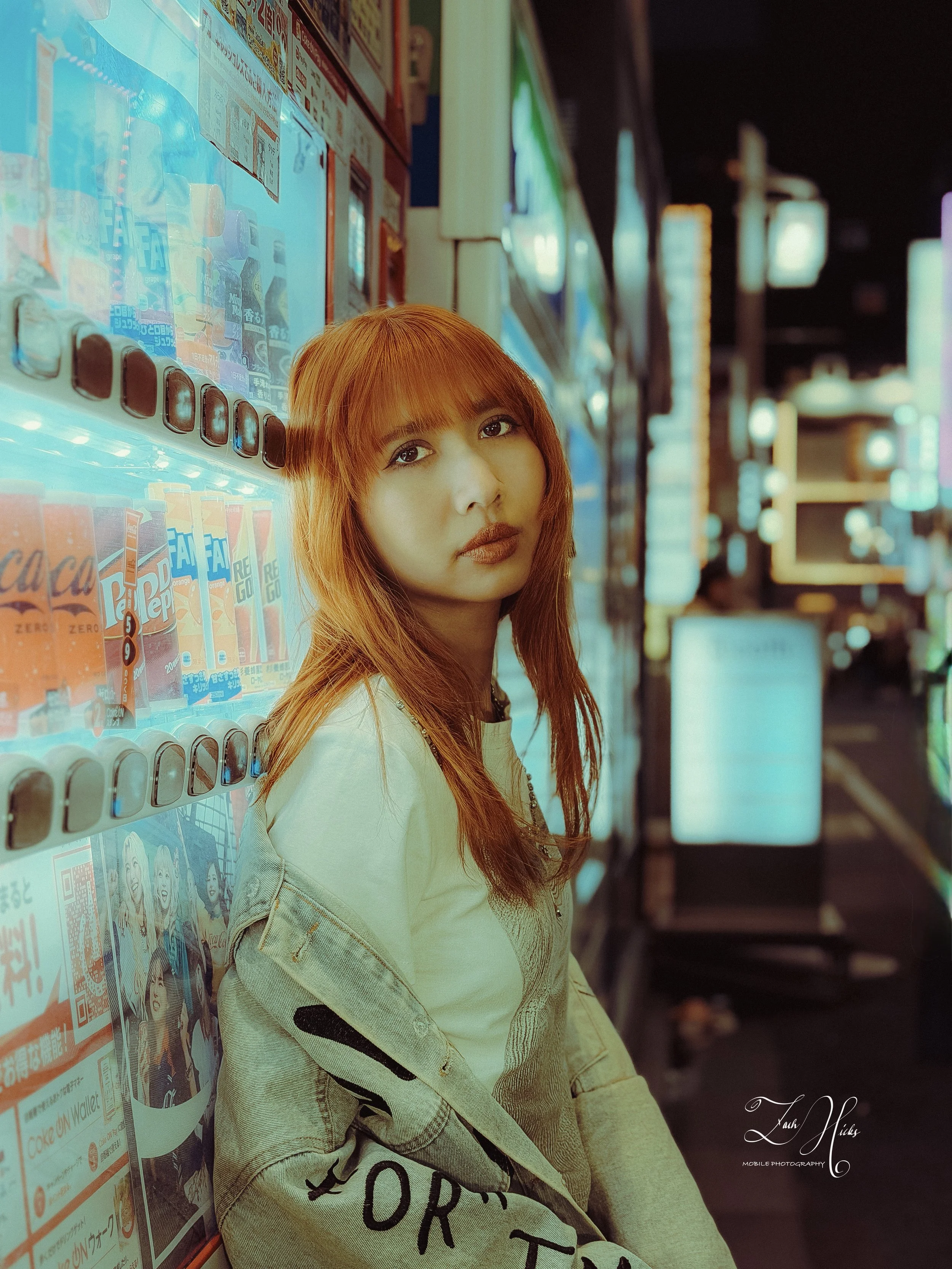 A young woman with reddish-brown hair and makeup, wearing a light-colored jacket, is standing next to a vending machine in a brightly lit urban setting at night. She is looking at the camera.