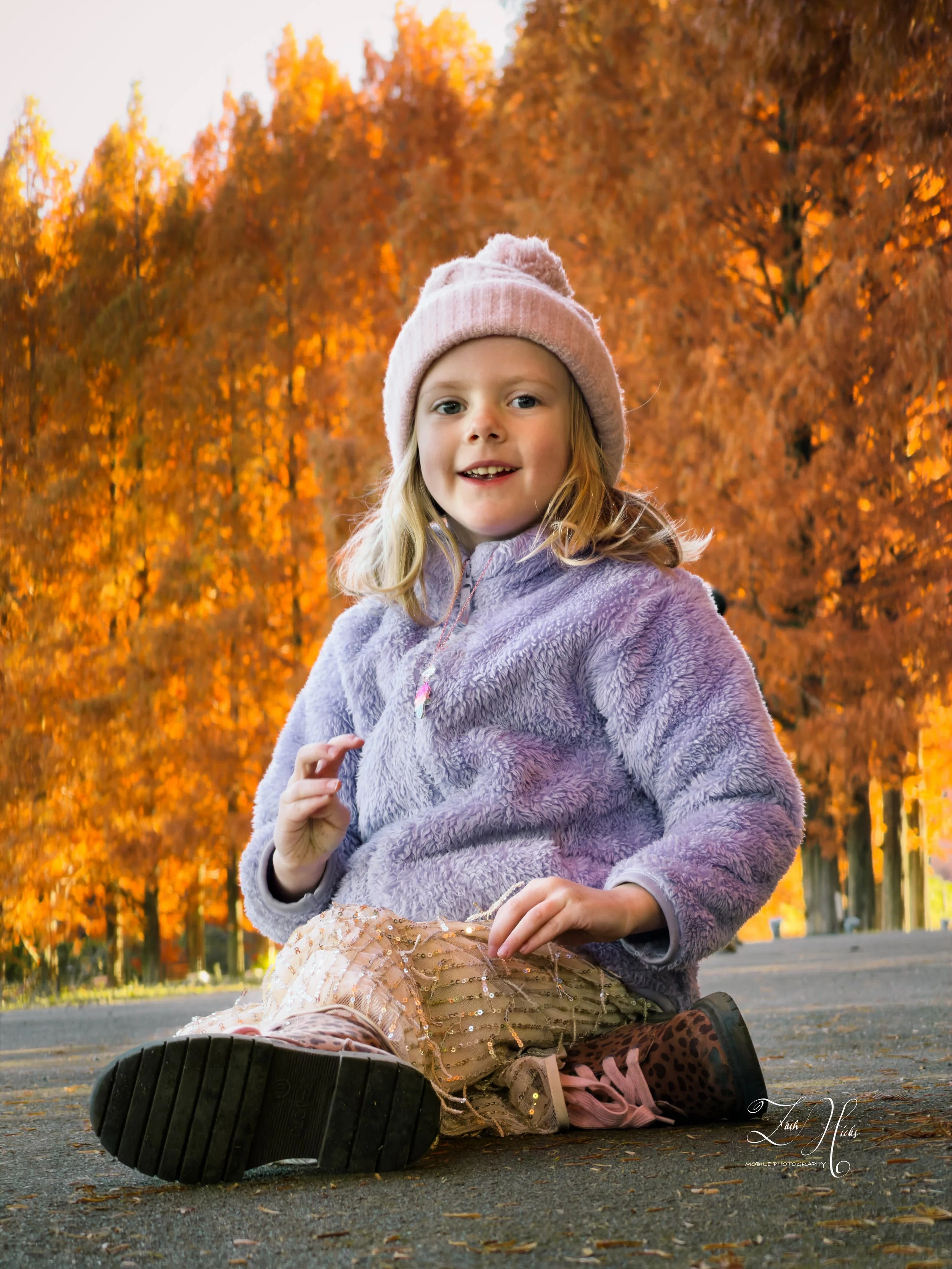 A young girl with blonde hair, wearing a pink knit hat, purple fleece jacket, and patterned pants, sitting on a paved path with vibrant autumn trees with orange leaves in the background.