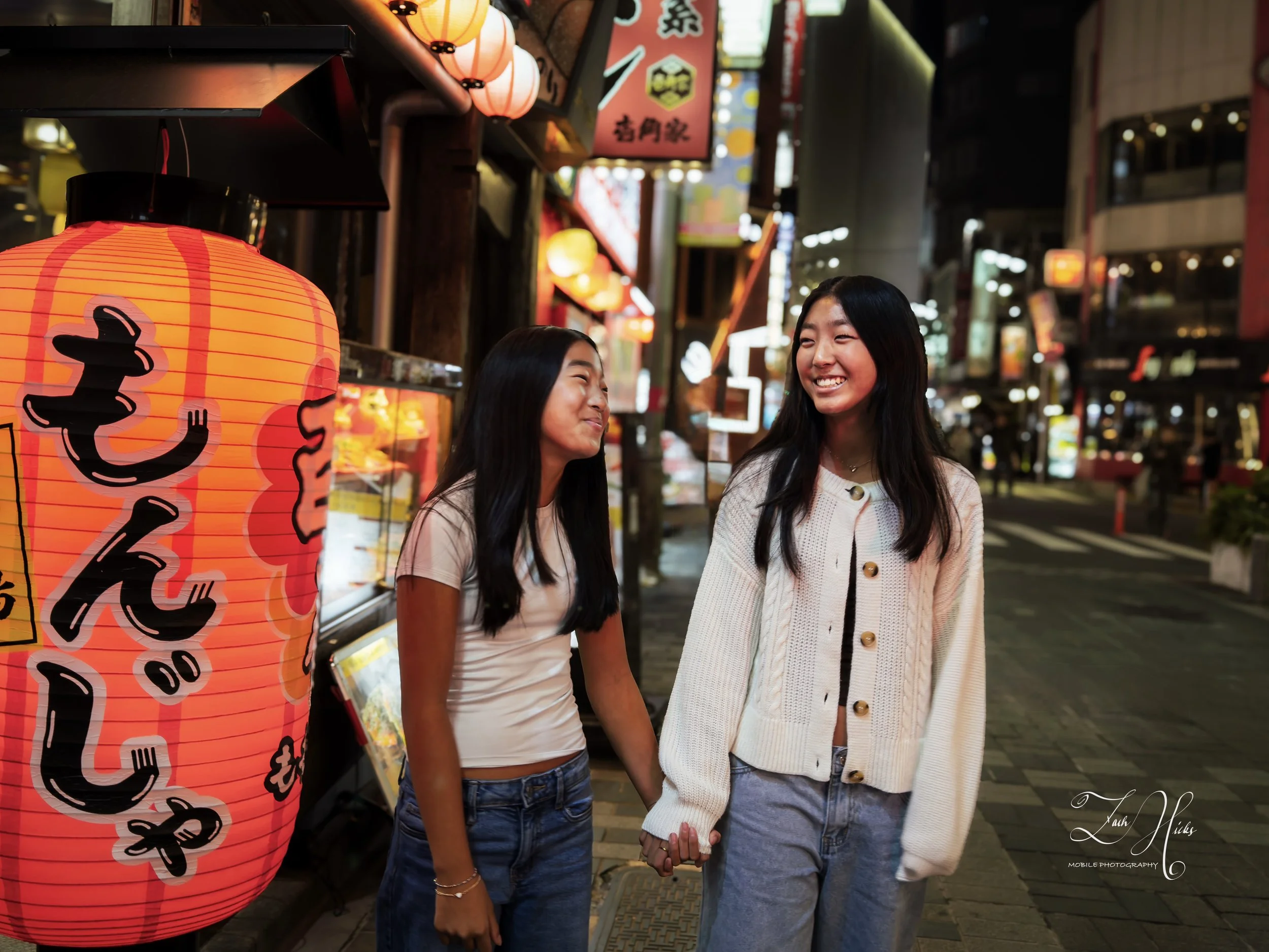 Two women holding hands and smiling on a vibrant night street in Japan, illuminated by red paper lanterns and surrounded by colorful signs and shopfronts.