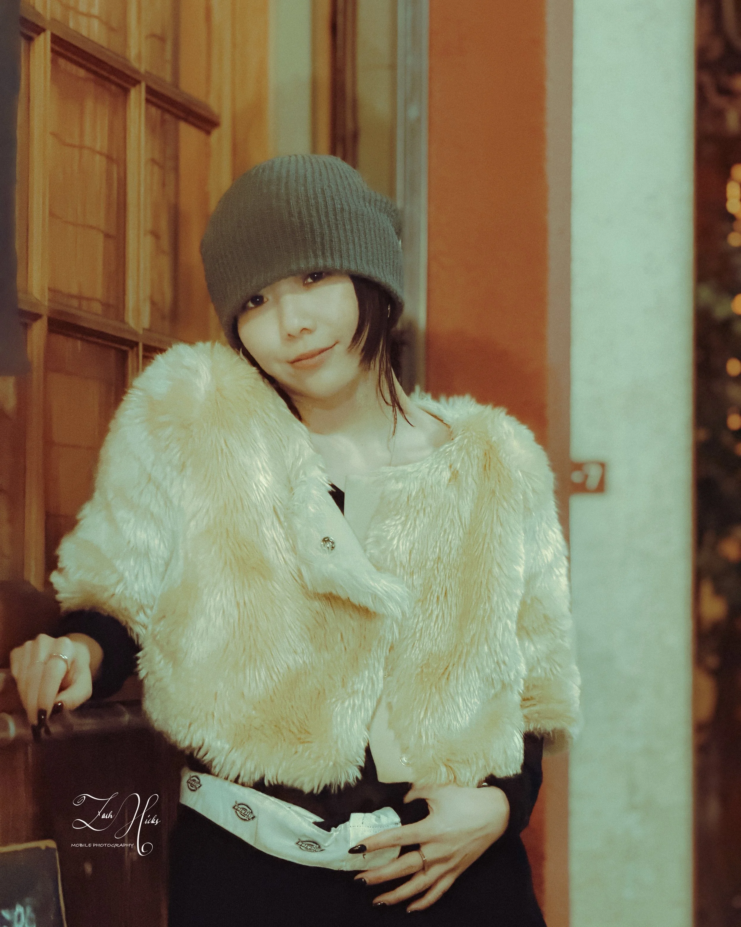 A woman with short black hair wearing a gray beanie, a cream-colored fluffy jacket, and black clothing underneath, standing indoors near a wood-paneled wall and a stone wall.