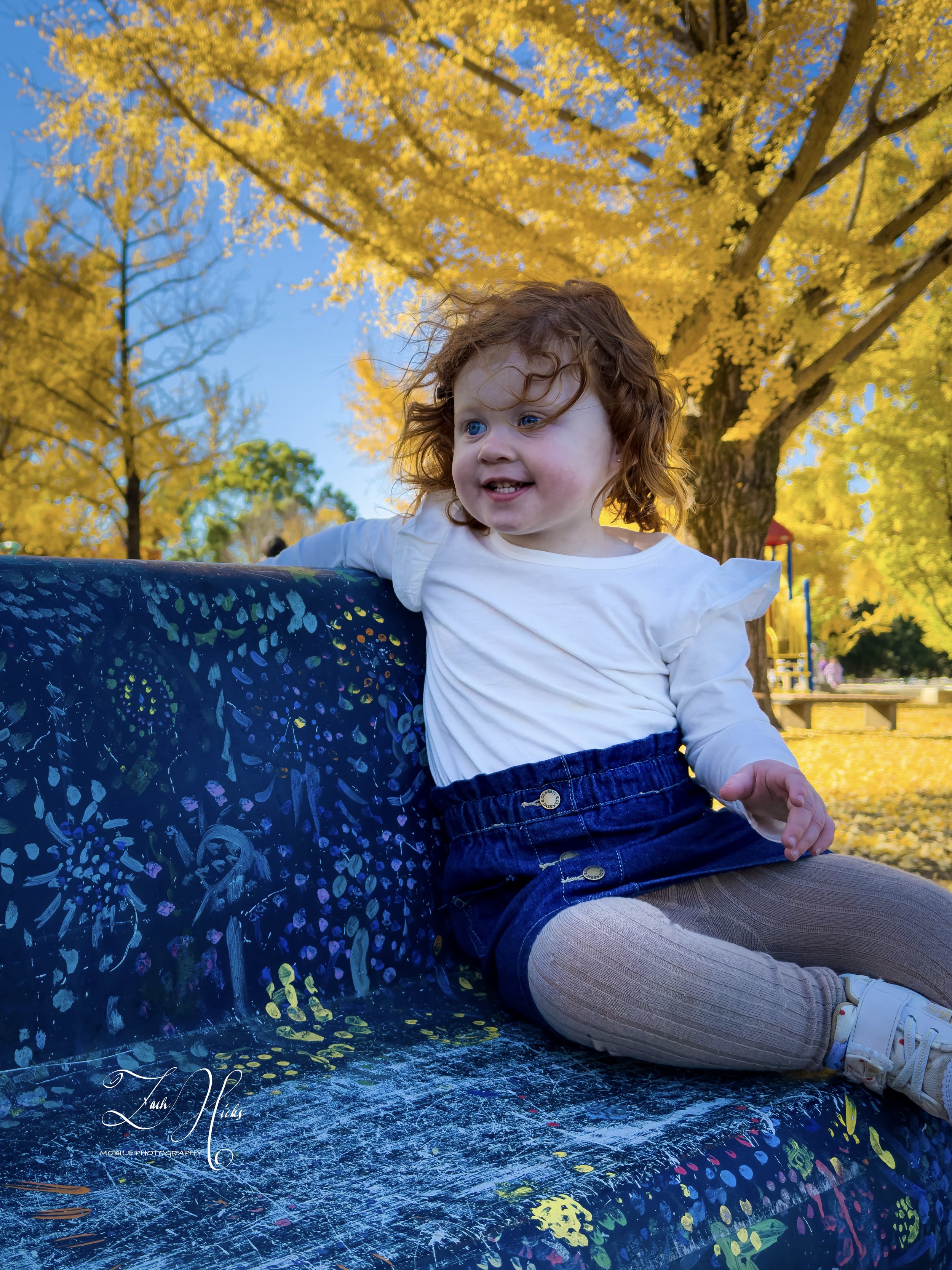A smiling young girl with curly red hair sitting on a painted park bench during fall, with yellow leaves and trees in the background.