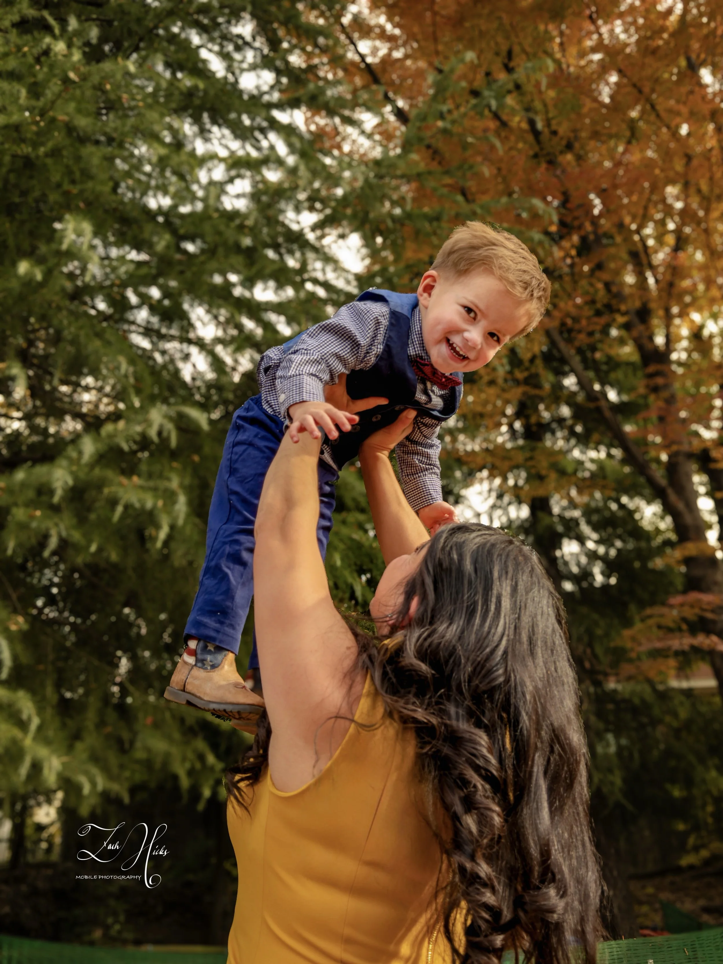 A woman with long dark curly hair, wearing a yellow sleeveless top, is lifting a young boy in a blue outfit, with a red bow tie, into the air outdoors. The background shows green and autumn-colored trees, indicating a fall setting.