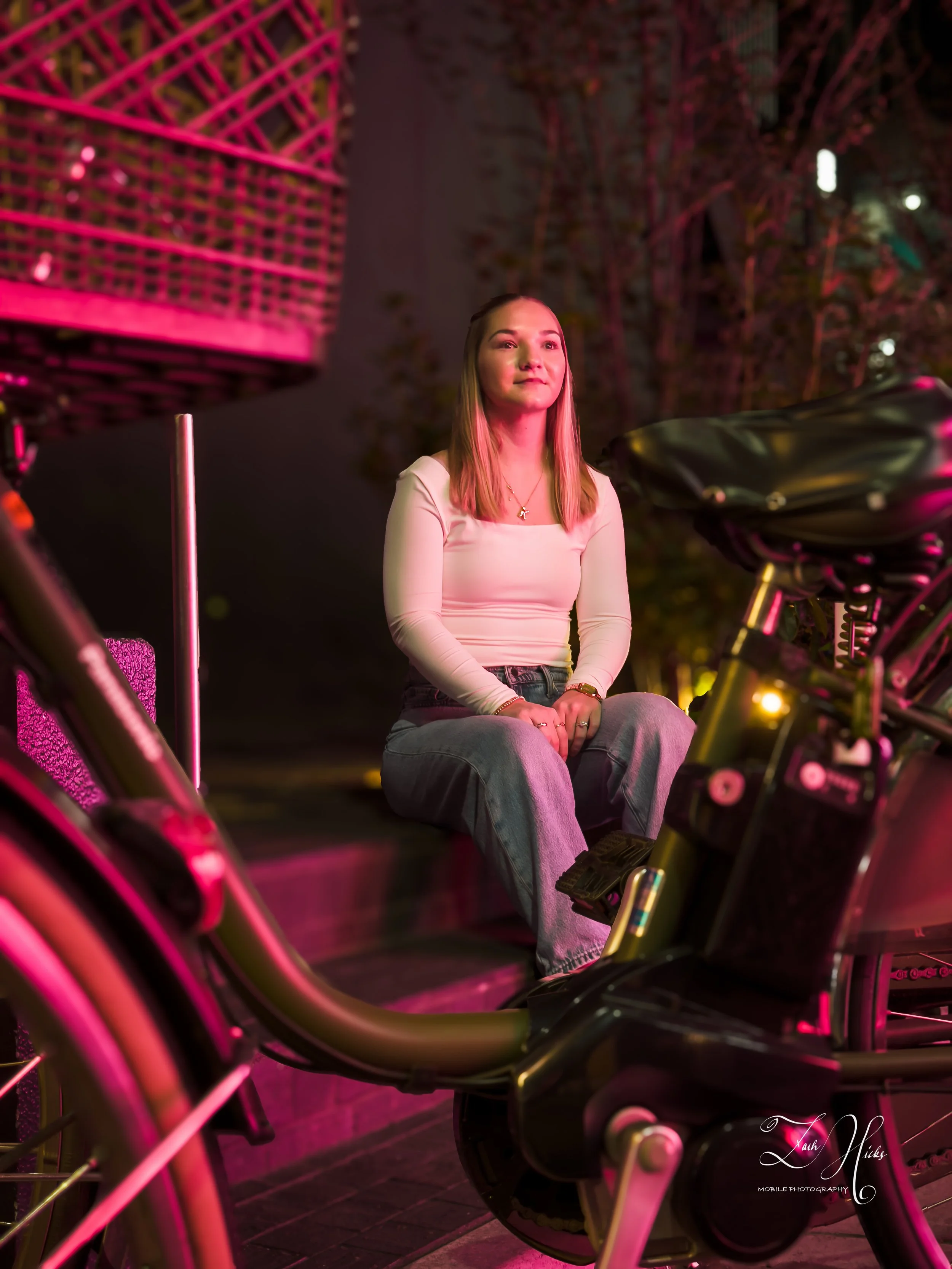 A woman sitting on a bicycle at night illuminated by pink and yellow lights, with trees in the background.