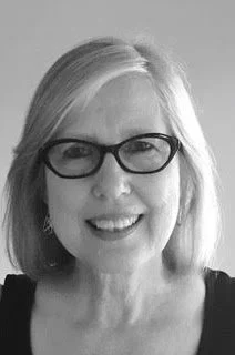 Portrait of a woman with blonde hair and glasses smiling in front of a light background.