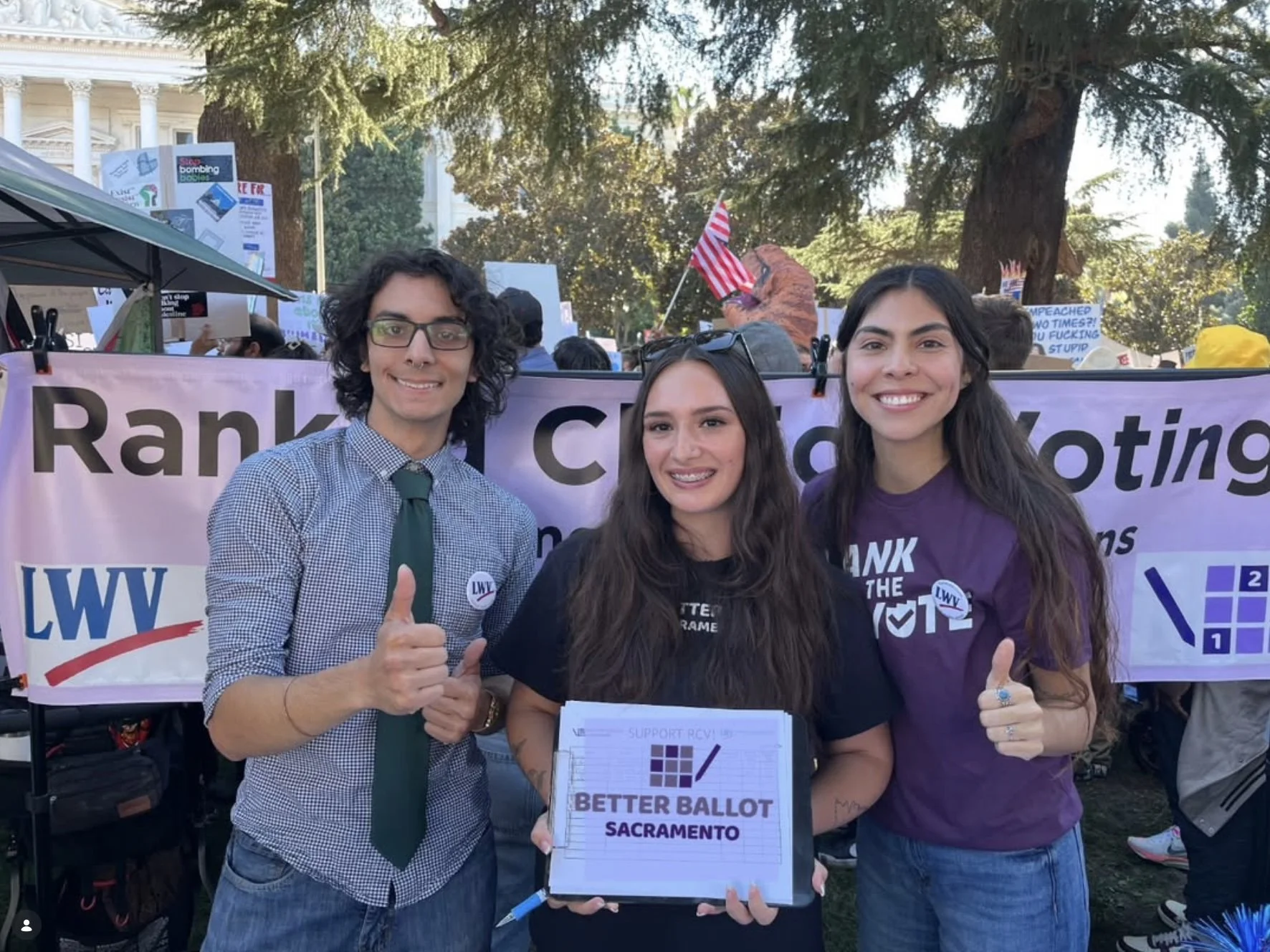 Three people standing in front of a protest sign that reads 'Ranked Choice Voting.' They are smiling, giving a thumbs up, and holding a sign that says 'Better Ballot Sacramento.' The group includes a man wearing glasses and a checkered shirt with a green tie, a woman with long dark hair in a black shirt, and a woman with long dark hair in a purple shirt. There are other protest signs and a crowd in the background.