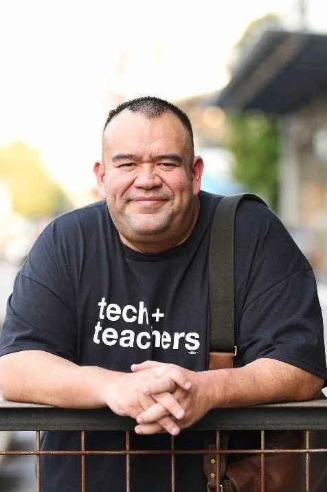 A man with short dark hair and a broad smile, wearing a black t-shirt that reads 'tech+teachers', leaning on a railing outdoors with trees and a building in the background.