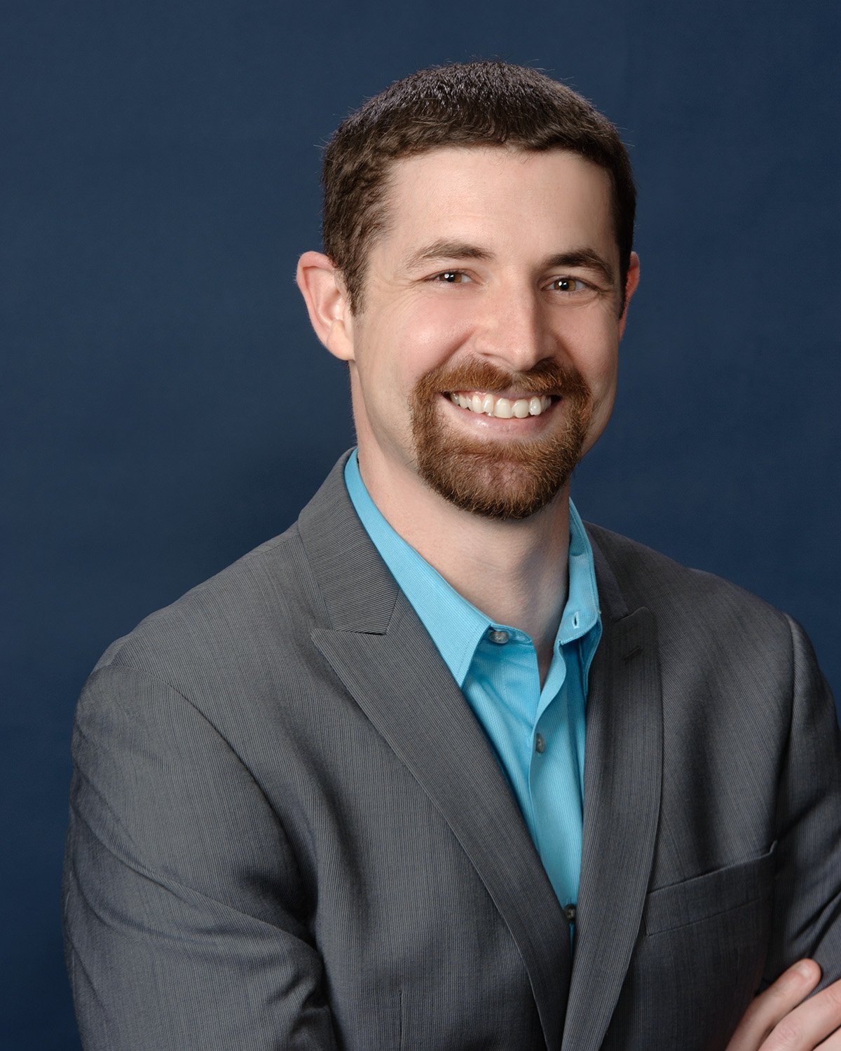 Headshot of a smiling man with a beard and mustache, wearing a gray suit and a light blue collared shirt, posed against a dark blue background.