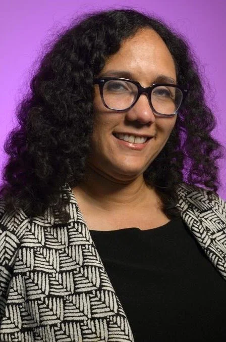 A woman with curly black hair and glasses smiling, wearing a black top and a black and white patterned blazer, against a purple background.