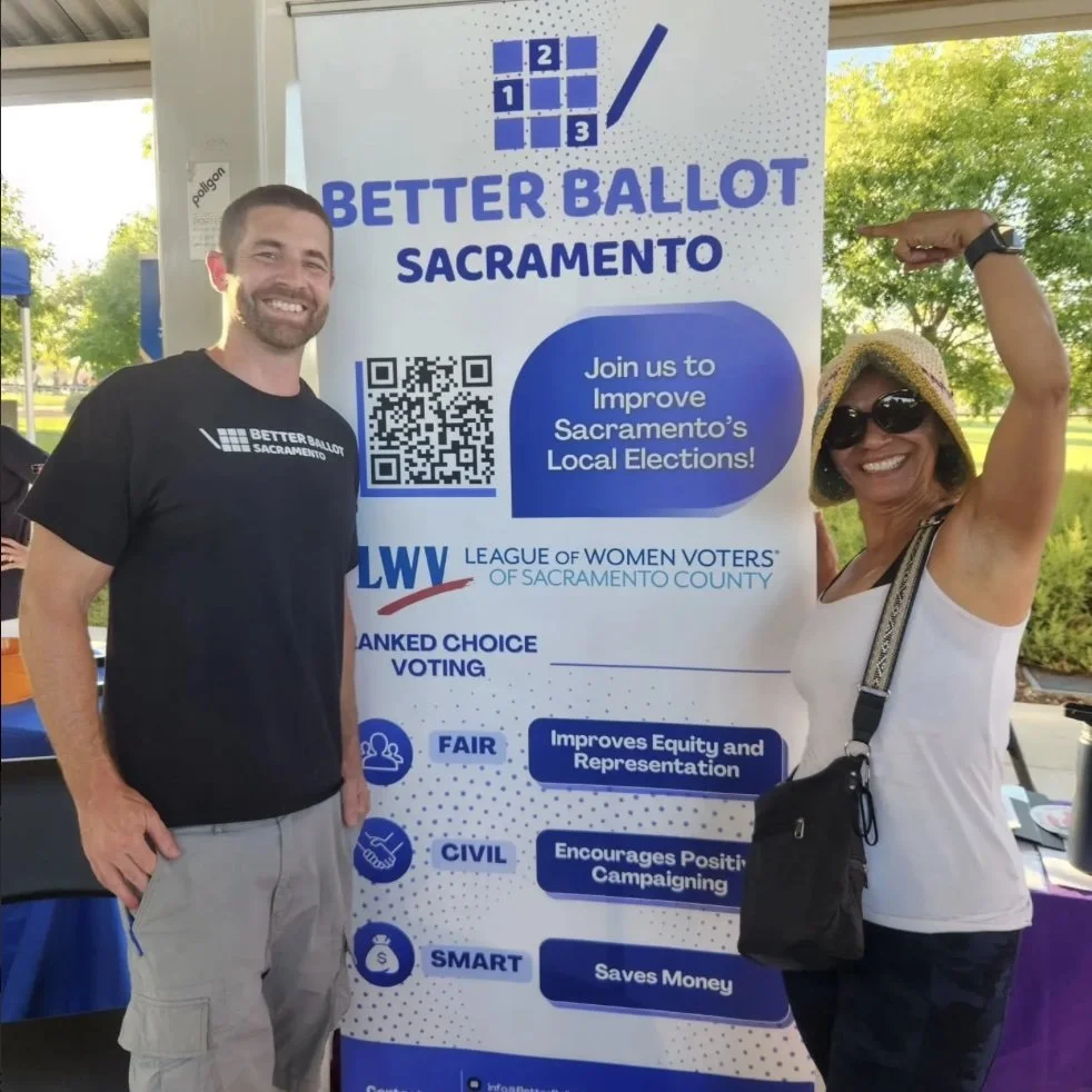 Two smiling women standing next to a large sign promoting Better Ballot Sacramento, a voting initiative. The woman on the right is pointing at the sign, wearing sunglasses and a sunhat, while the man on the left wears a black T-shirt with the same logo.