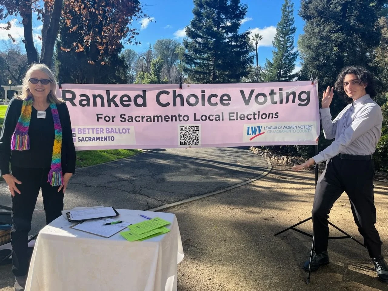 Two women standing outdoors near a large pink banner that reads "Ranked Choice Voting for Sacramento Local Elections." One woman on the left is wearing sunglasses, a black blazer, and a colorful scarf, and is standing next to a table with papers and green brochures. The other woman on the right is wearing a white shirt and black trousers, and is adjusting a pole or stand holding the banner.