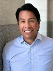 Portrait of a smiling man wearing a light blue dress shirt, standing against a neutral background.