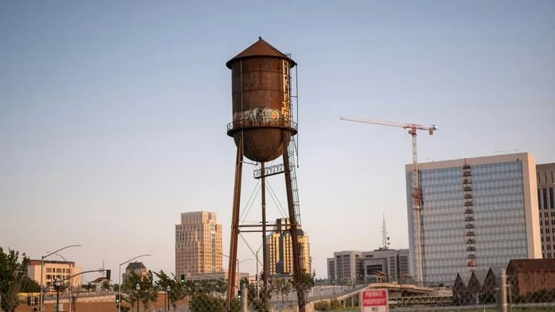 Old rusted water tower in an urban area with modern high-rise buildings and a construction crane in the background.